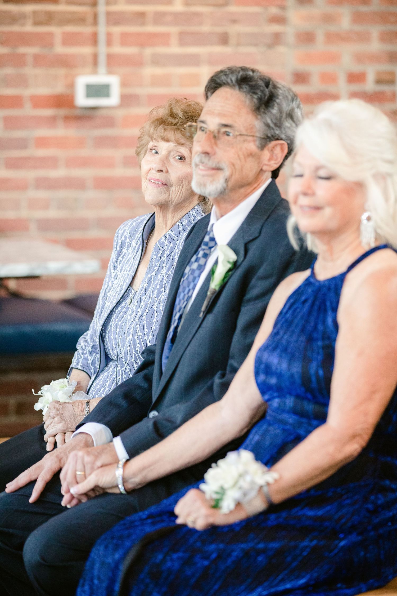 Guests seated in formal attire at a wedding ceremony with brick wall backdrop.