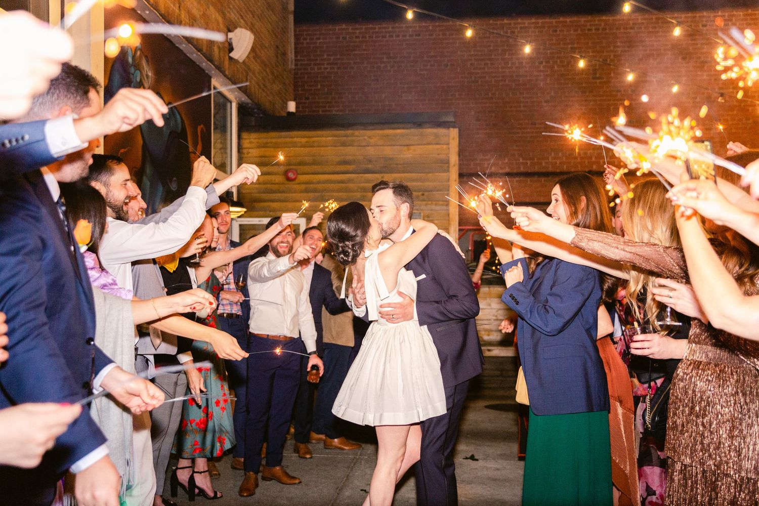 Wedding guests hold sparklers creating a magical nighttime sendoff tunnel outside a rustic venue.