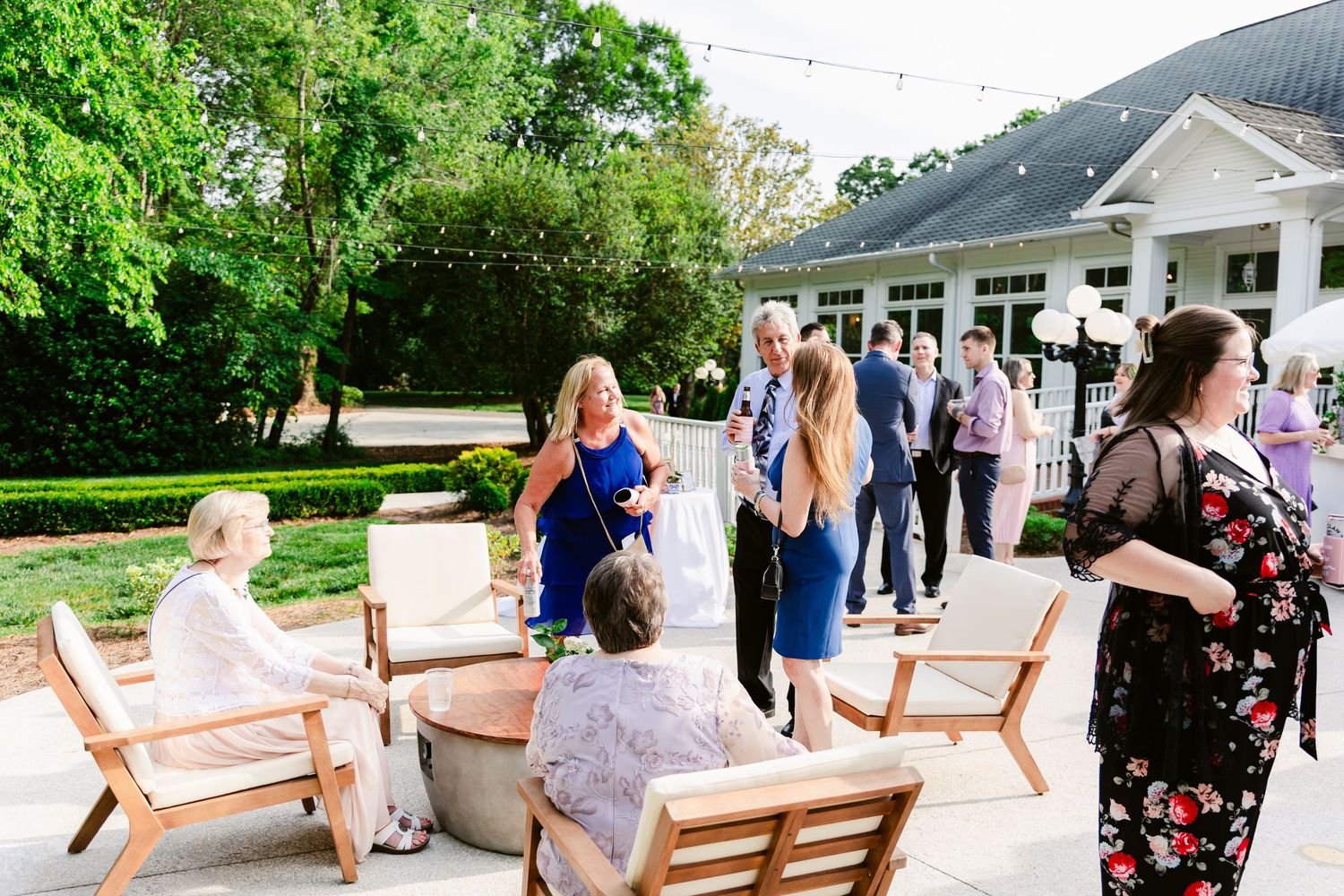 People gather on a white porch with wooden chairs during a social event at an elegant white house surrounded by greenery.