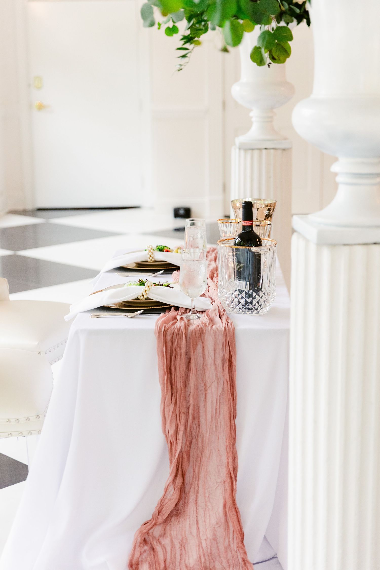 Elegant wedding reception tables with white linens and pink chiffon runners in a bright, airy venue with green foliage accents.