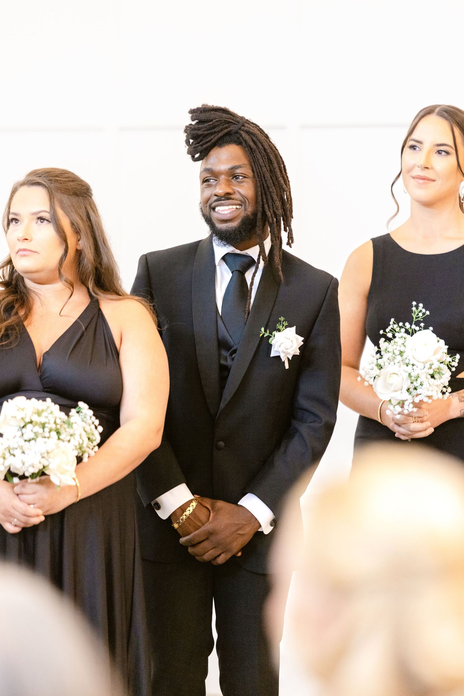Wedding party members in black attire and white dresses pose together holding white floral bouquets.