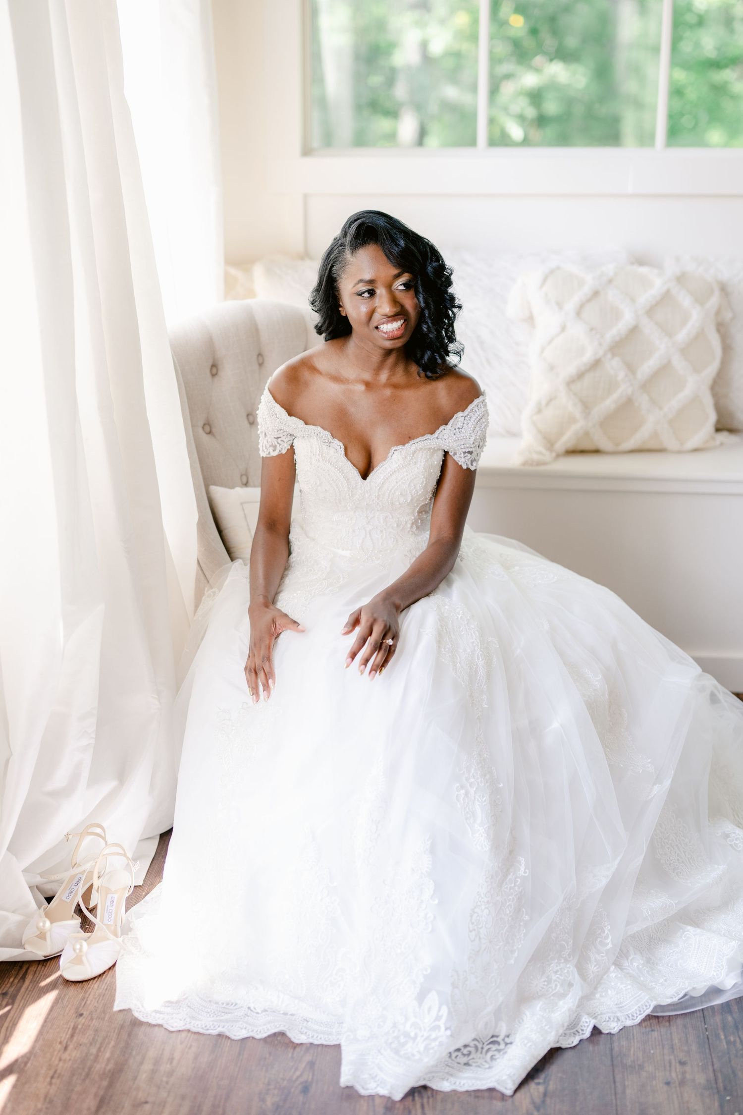 A stunning bride in a white lace wedding gown sits elegantly on a white sofa in a bright sunlit room.