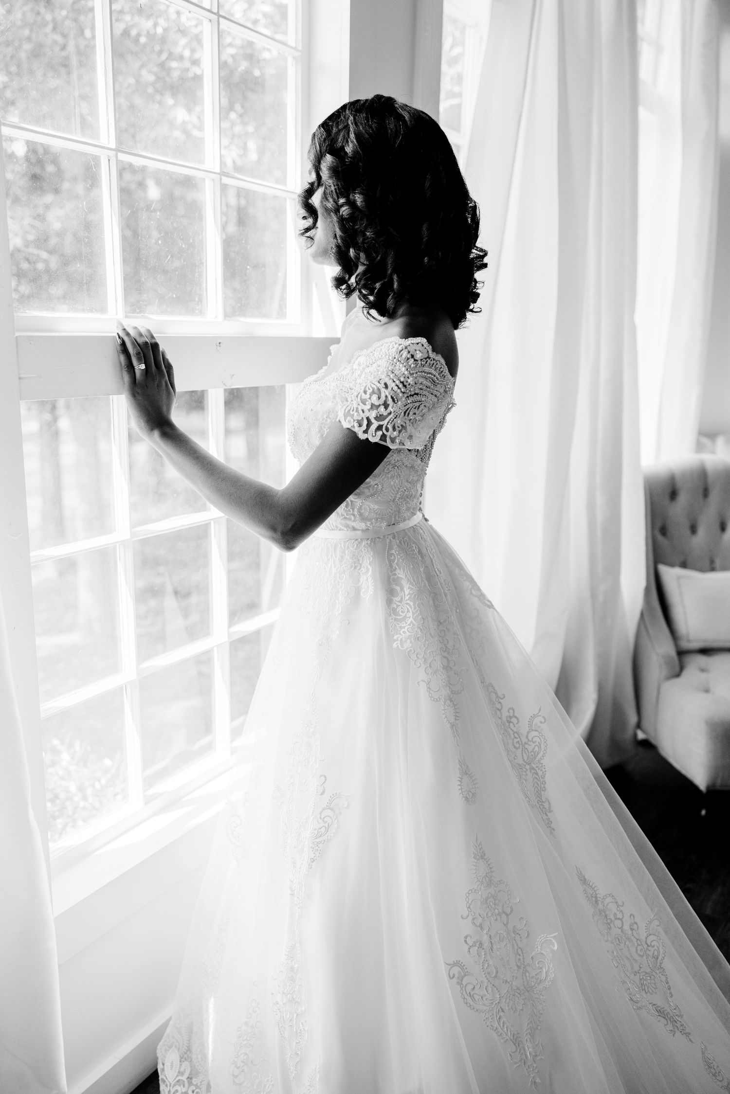 Black and white silhouette of a bride in a lace wedding gown with curled hair standing by a sunlit window.