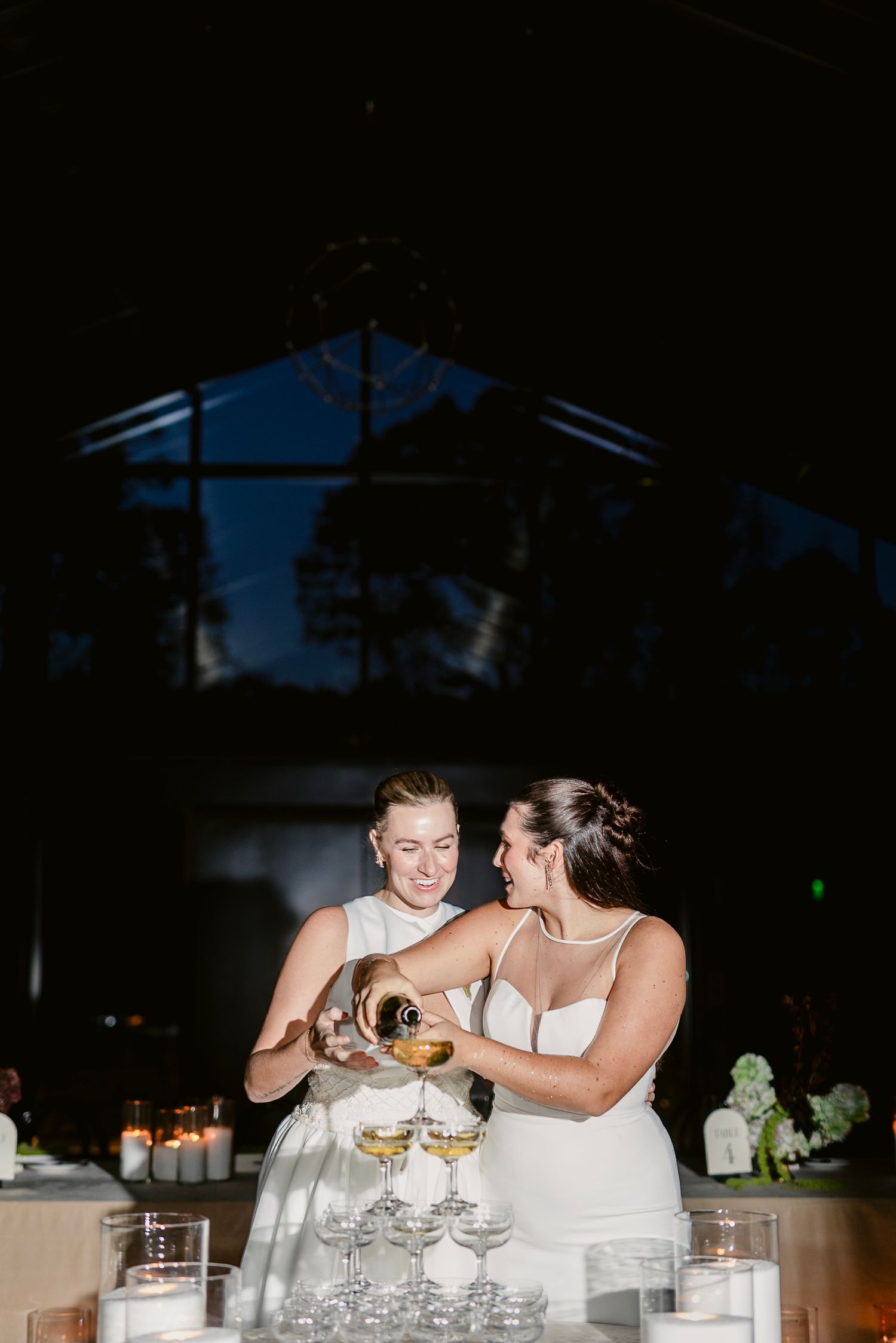 Two brides in white dresses share a toast and laugh together at their wedding reception.