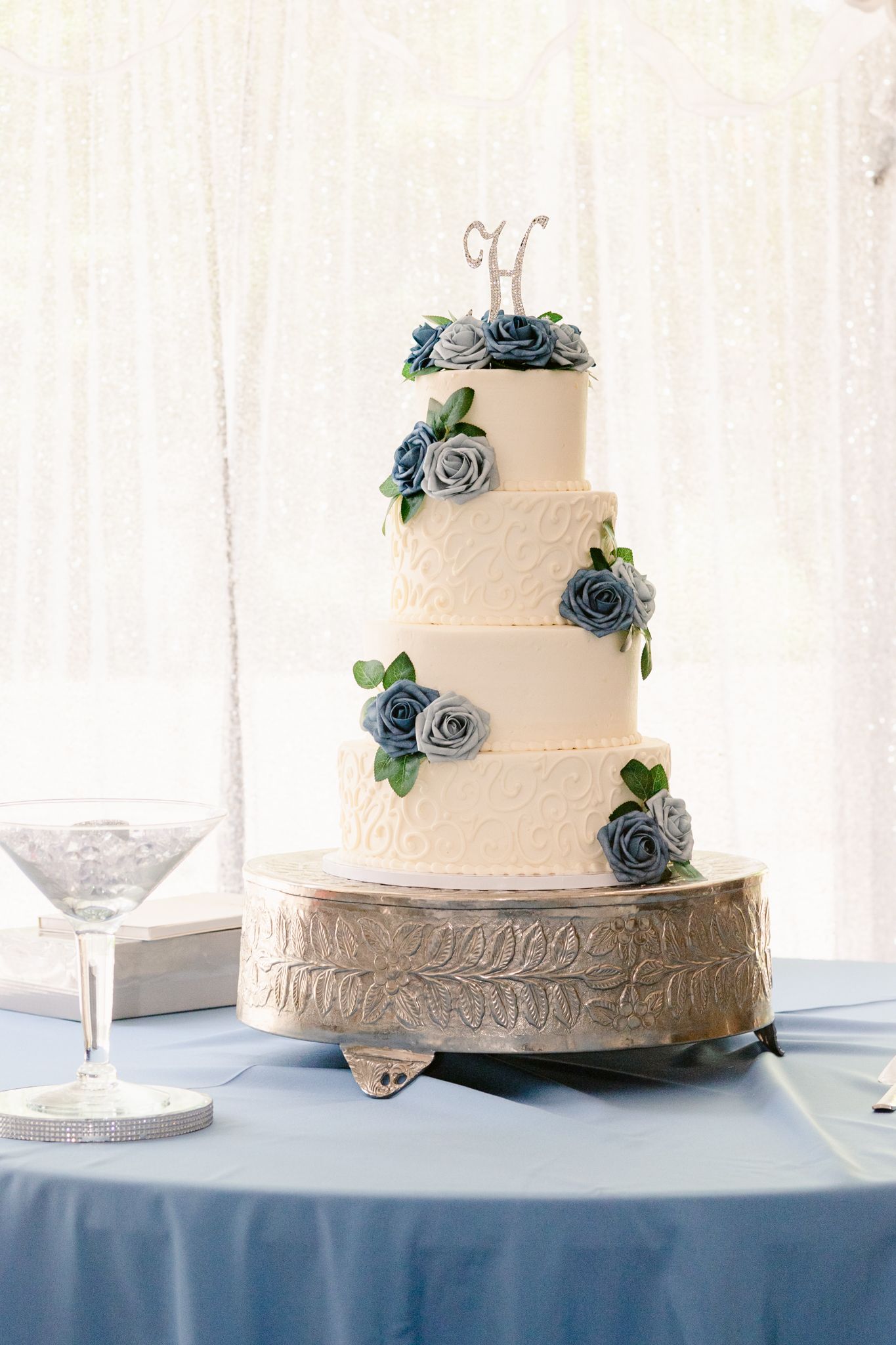 White tiered wedding cake decorated with dusty blue roses and delicate piping on a silver cake stand.