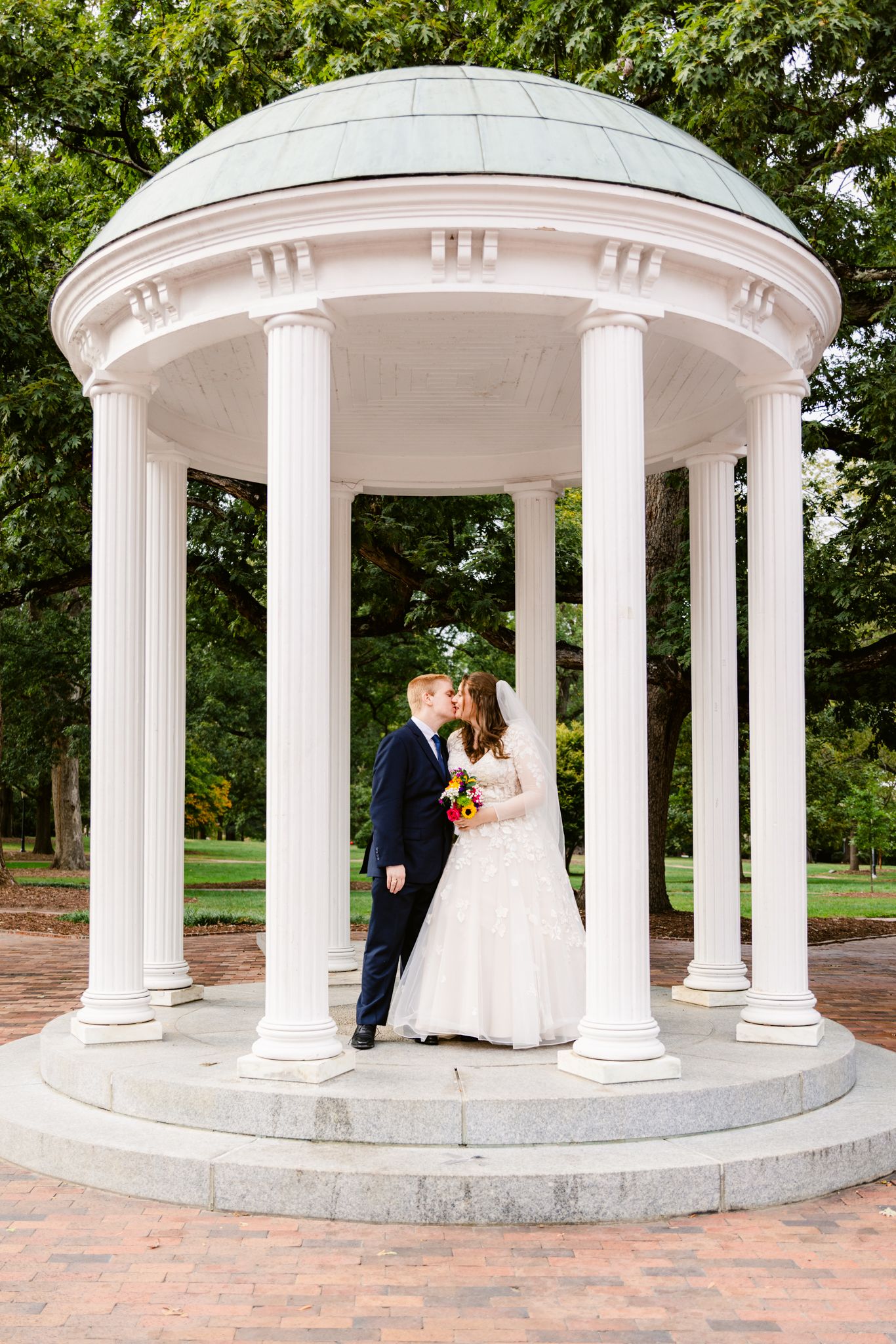 A romantic couple poses in a series of elegant white gazebo photos at a park with brick pathways and lush greenery.