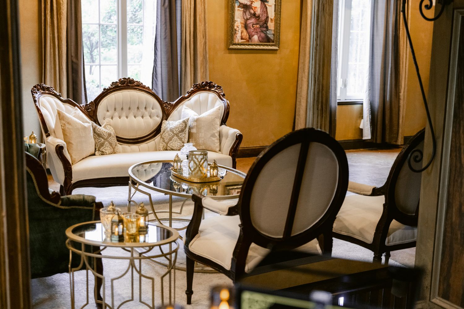 Elegant restaurant interior with tufted cream chairs and dark wood tables arranged beneath ornate gold walls and chandeliers.