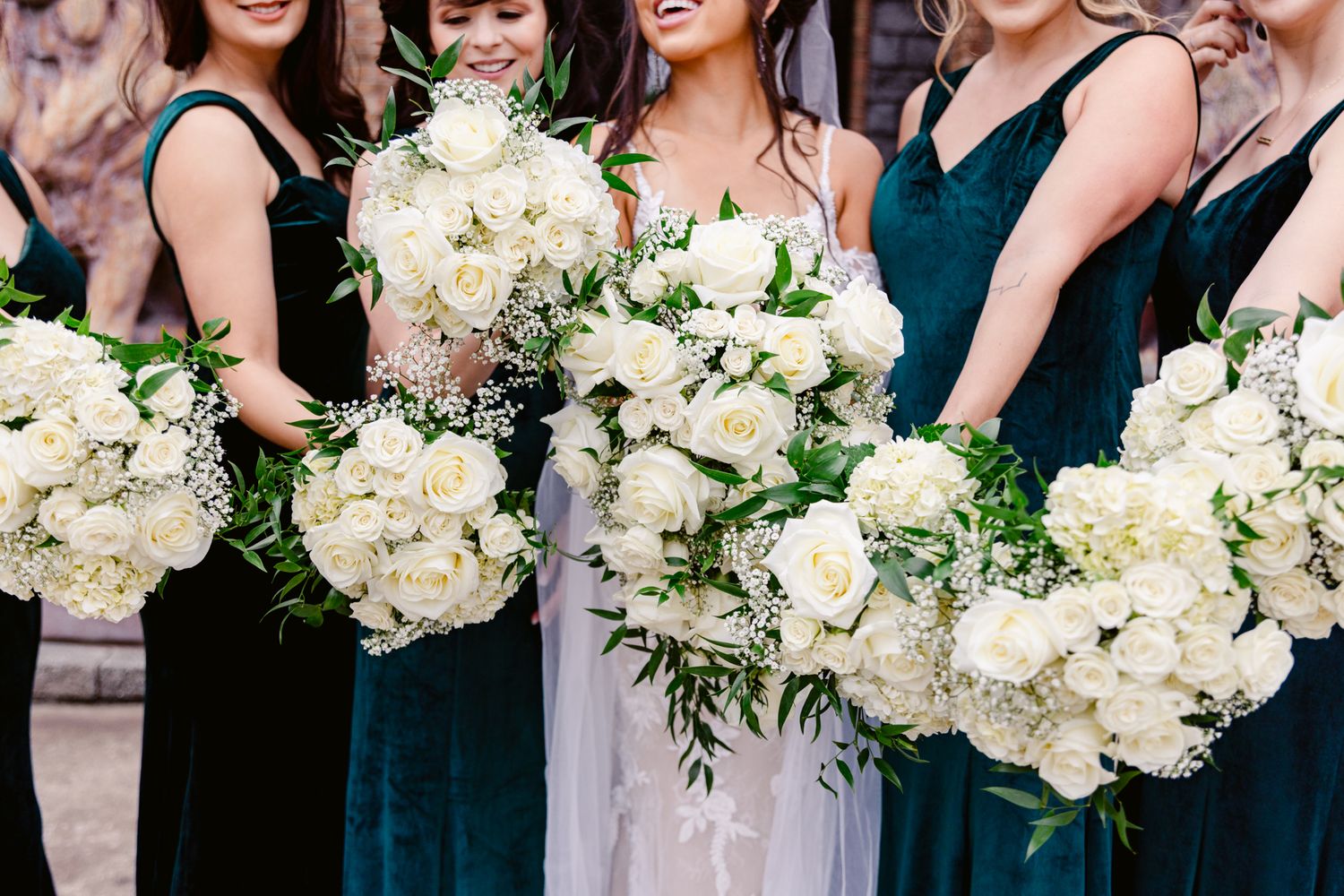 Wedding party members hold elegant white floral bouquets while wearing dark teal dresses at an outdoor celebration.