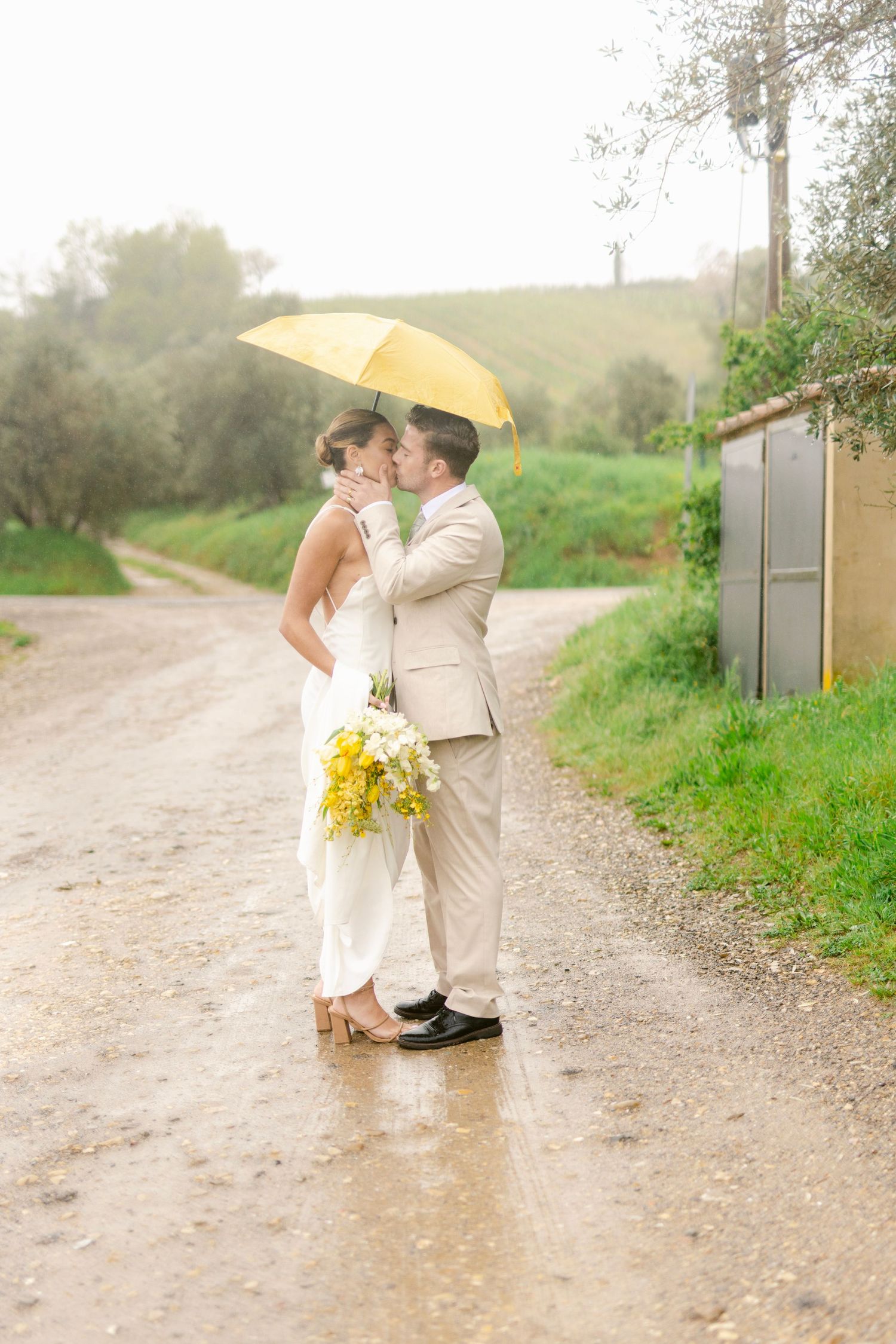 A couple shares a romantic moment under a yellow umbrella on a misty country path.