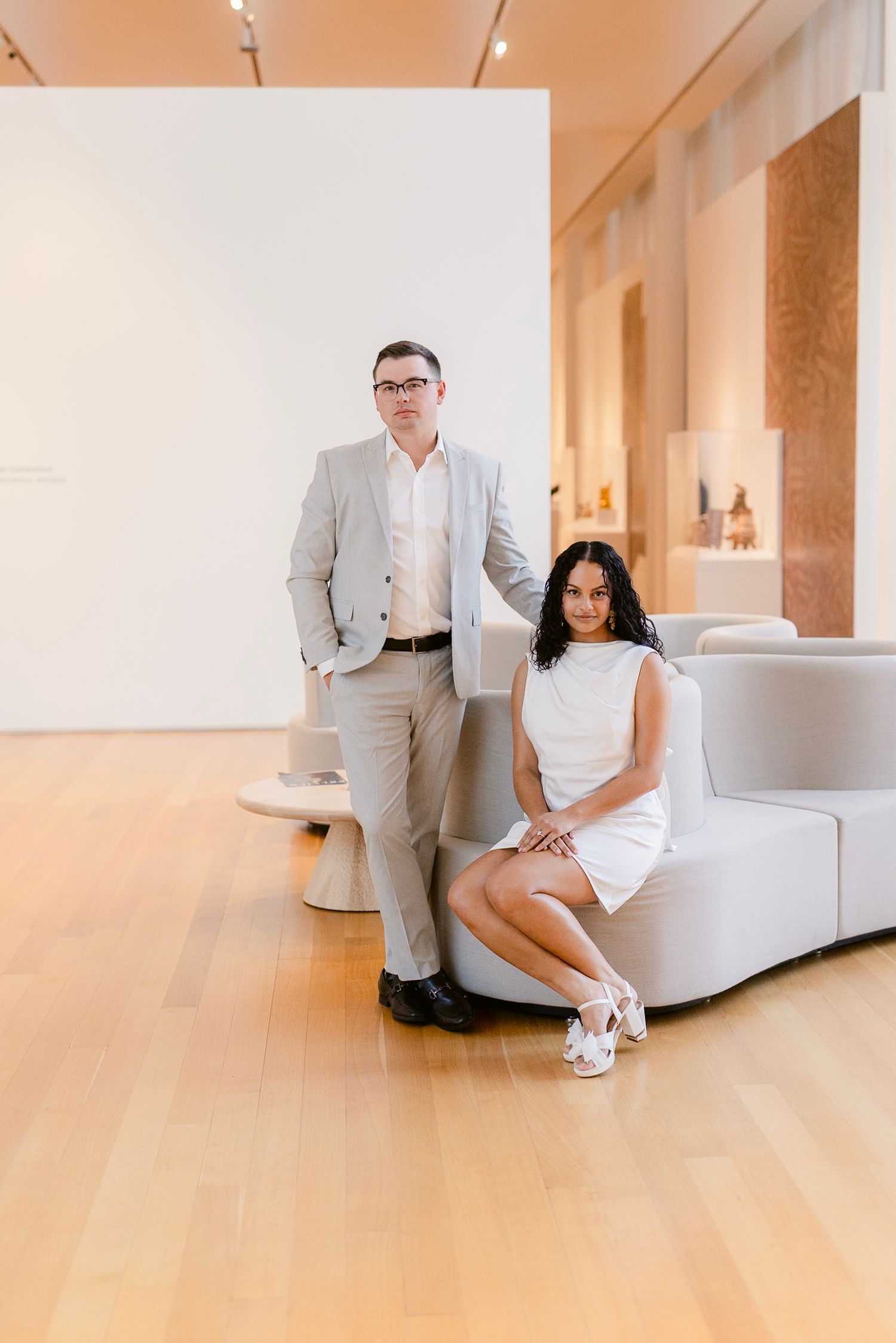 A couple in coordinated light outfits pose together in a modern minimalist gallery space with hardwood floors.
