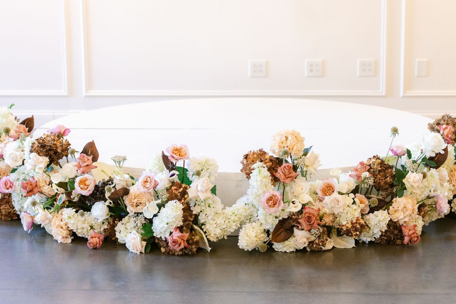 A curved floral arrangement of peach, cream and white roses with hydrangeas displayed on a dark floor.