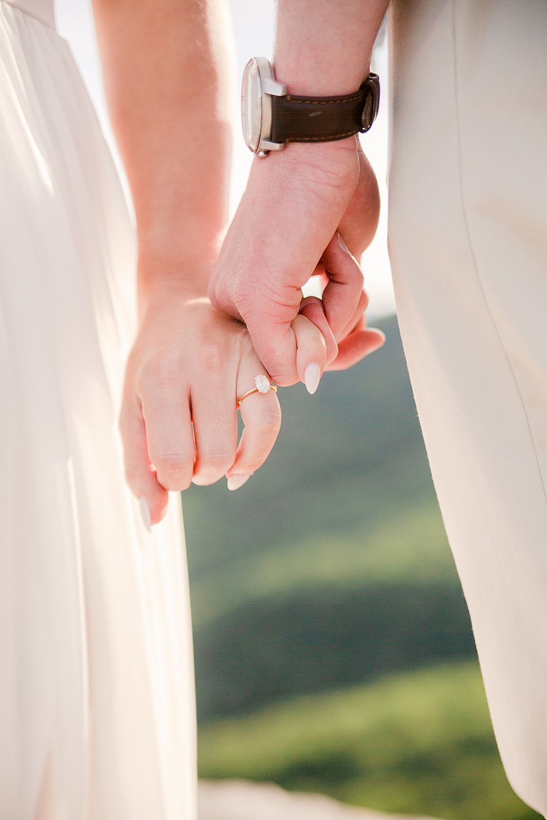 Two hands gently intertwined with one wearing a watch, set against a soft natural outdoor backdrop.