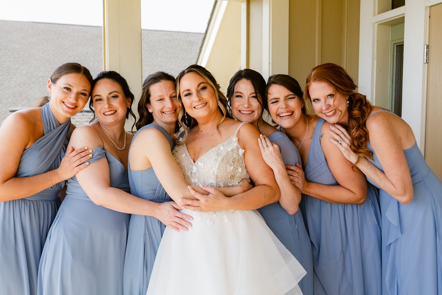 Bridesmaids in dusty blue dresses pose together for wedding photos in a bright indoor setting.