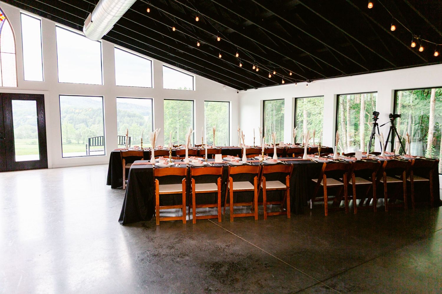 A long rustic reception hall with exposed wooden beams, string lights, and windows overlooking greenery outside.