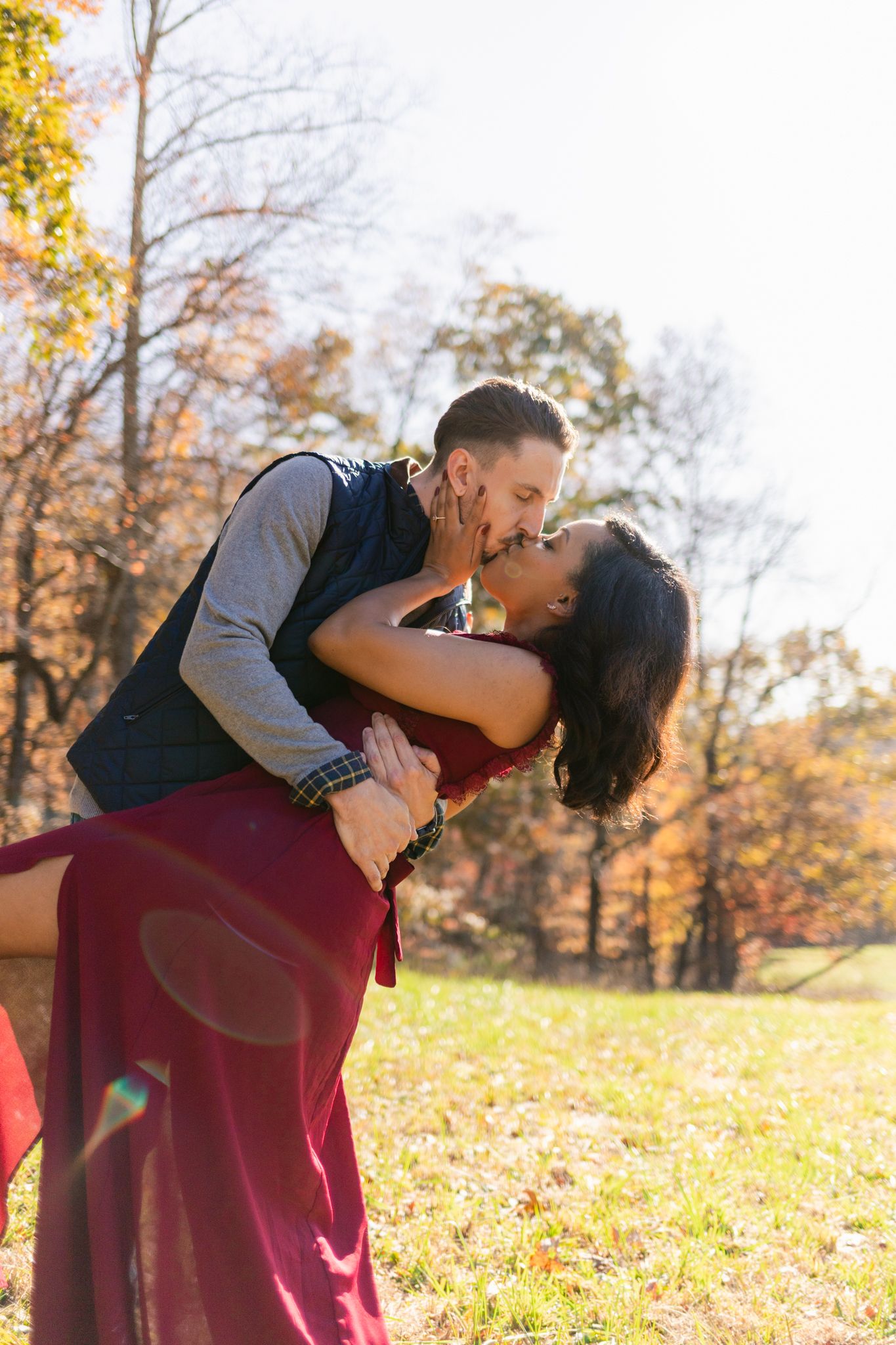A romantic couple embraces and kisses in a park filled with golden autumn leaves on a sunny fall day.