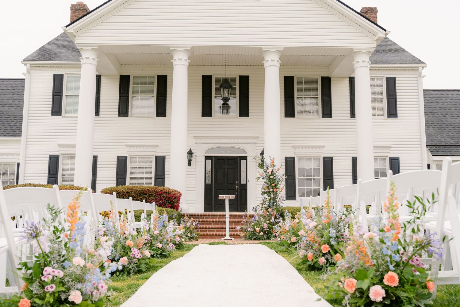 A white colonial mansion with grand columns features a symmetrical facade and colorful flower gardens lining the walkway.