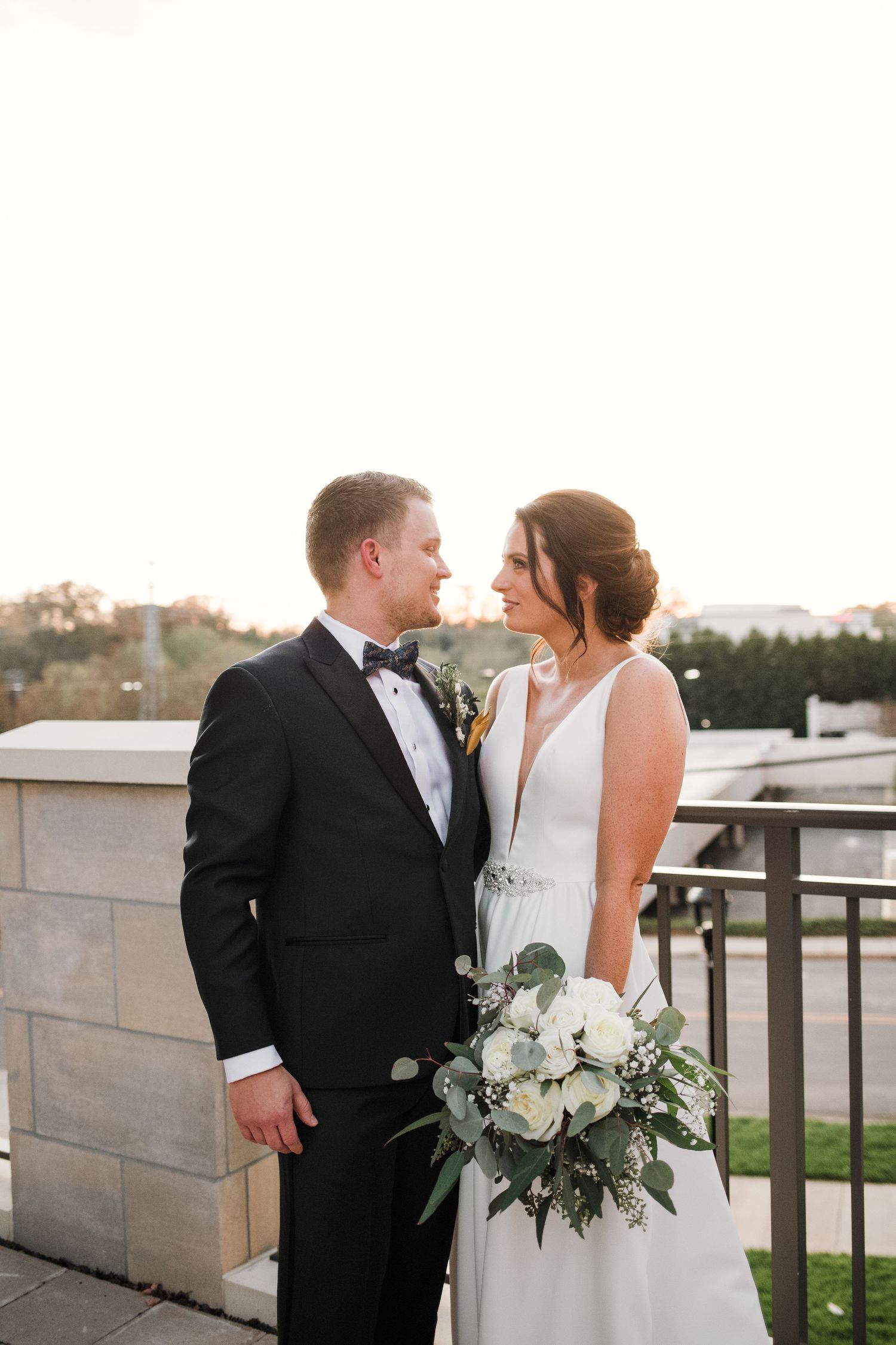 A romantic wedding portrait showing a couple on a rooftop terrace with a white floral bouquet at sunset.