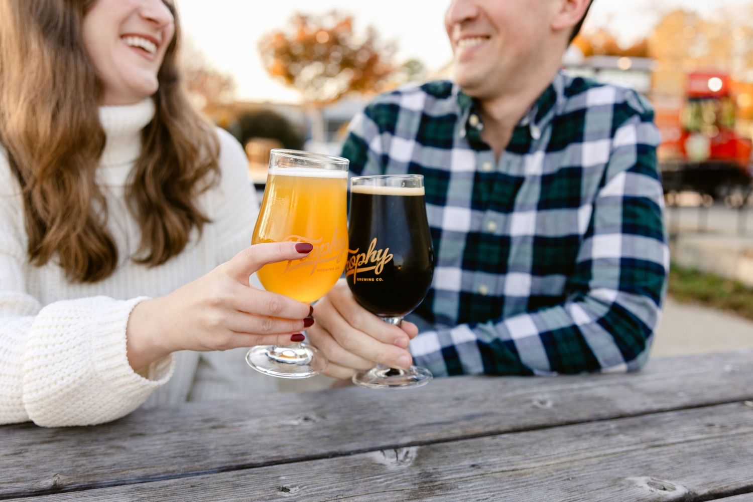 Two people share craft beer glasses while sitting at an outdoor wooden table in casual attire.