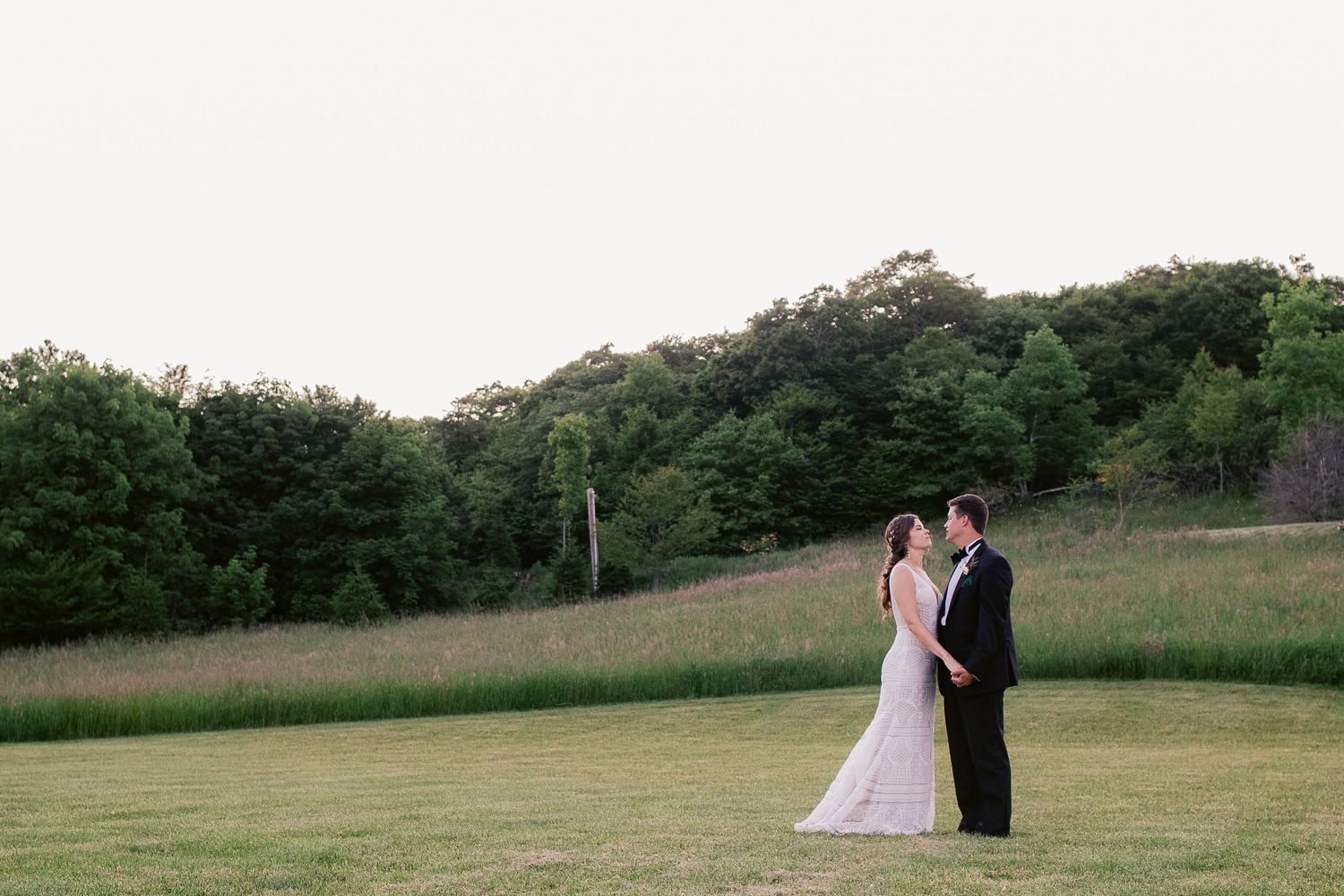 Wedding couple stands in open field with tree line and sunset backdrop.