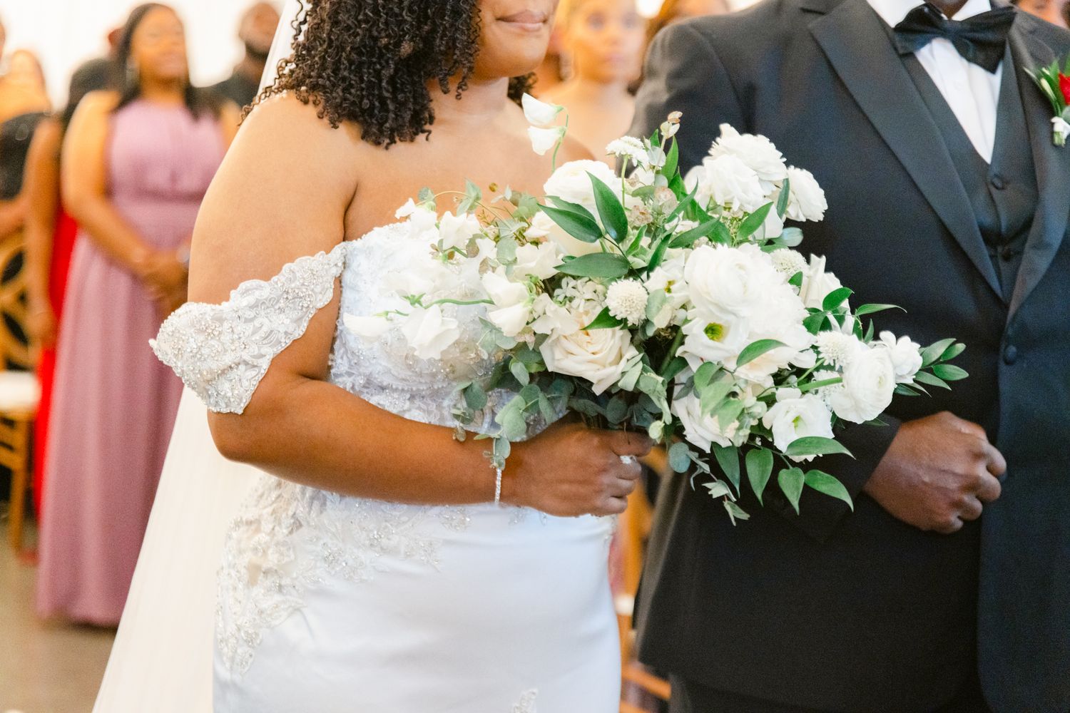 Beautiful white bridal bouquet with lilies and greenery held during wedding ceremony next to black tuxedo.