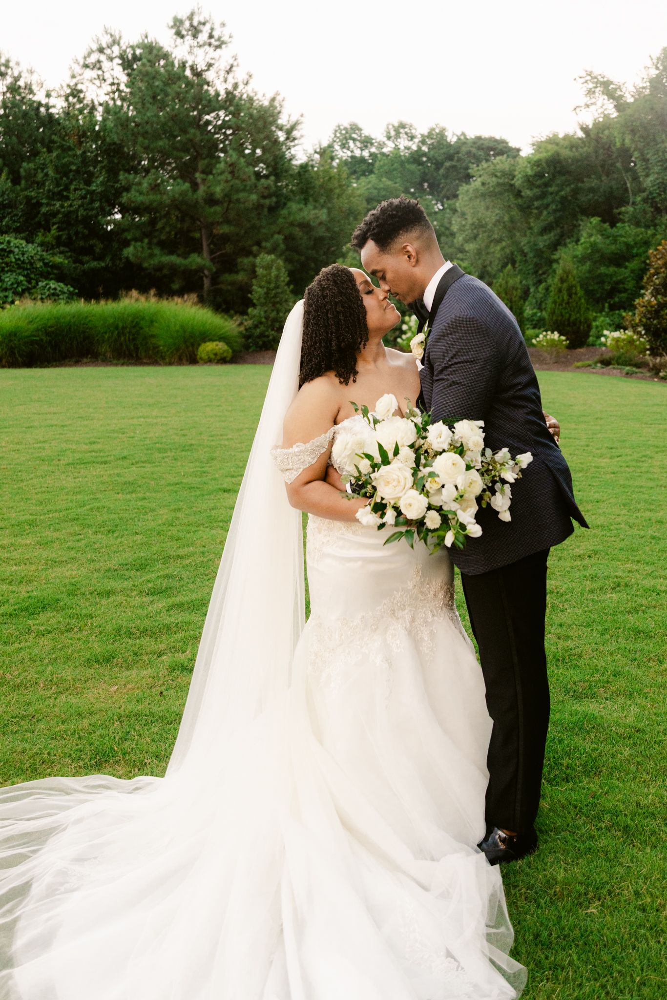 A romantic wedding moment captured on a lush green lawn with a beautiful white bouquet and flowing veil.