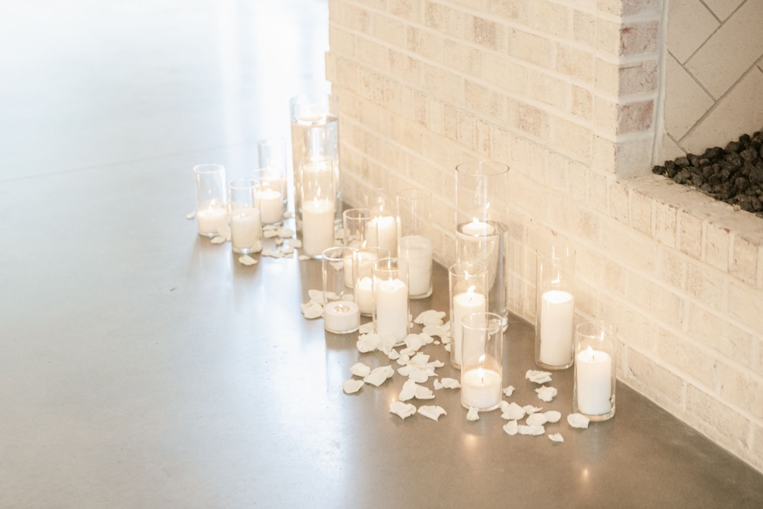 Row of glowing white pillar candles with scattered petals creating romantic ambiance along a wall.