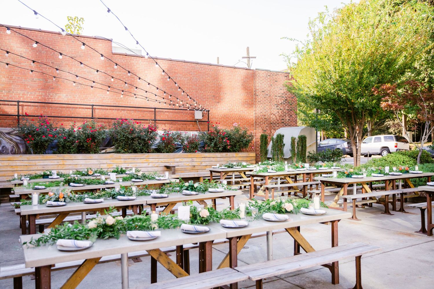 A panoramic view of outdoor picnic tables and benches arranged in rows next to a brick building and garden area.