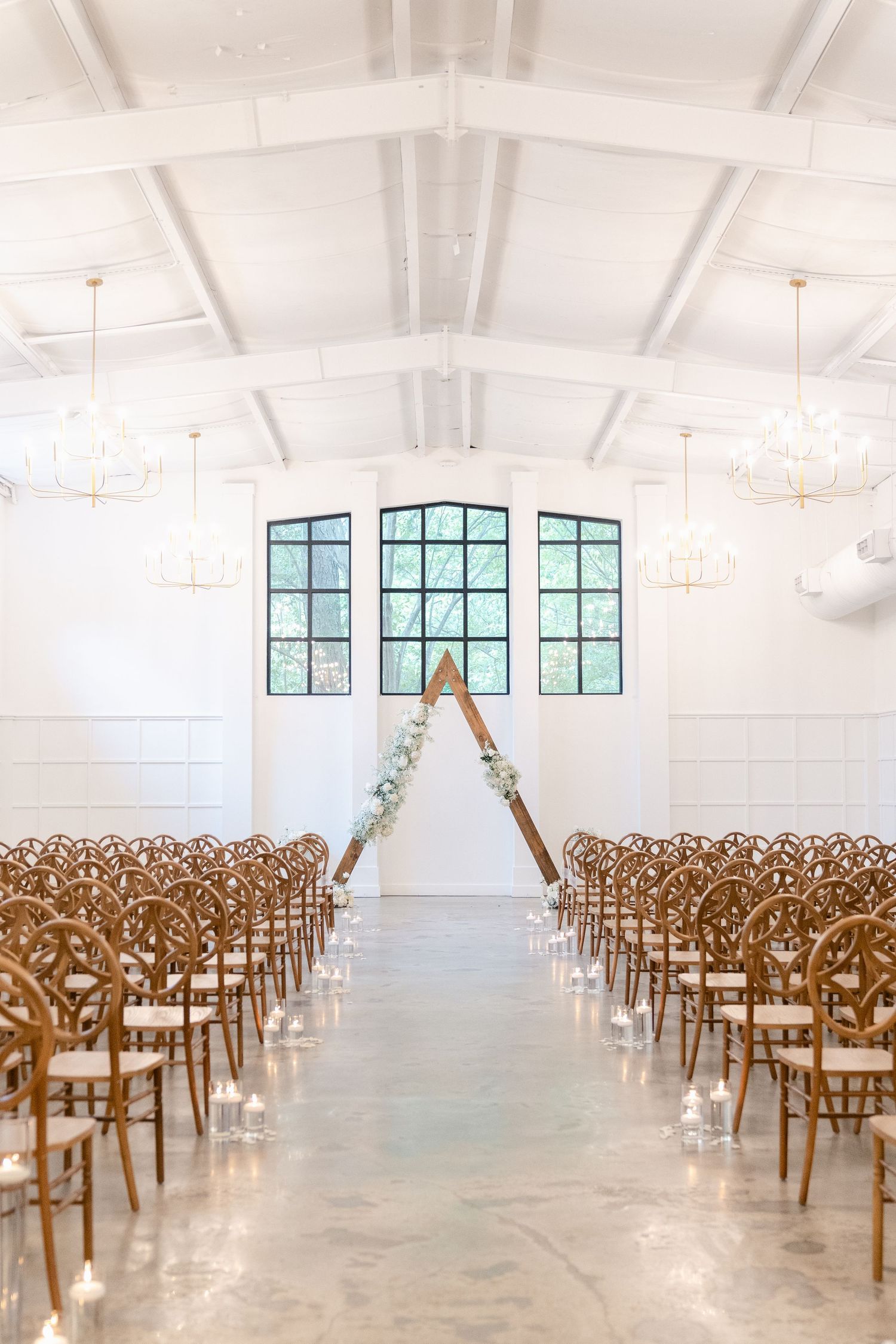 Wedding ceremony space with wooden cross-back chairs and geometric wooden arch in a bright industrial venue.