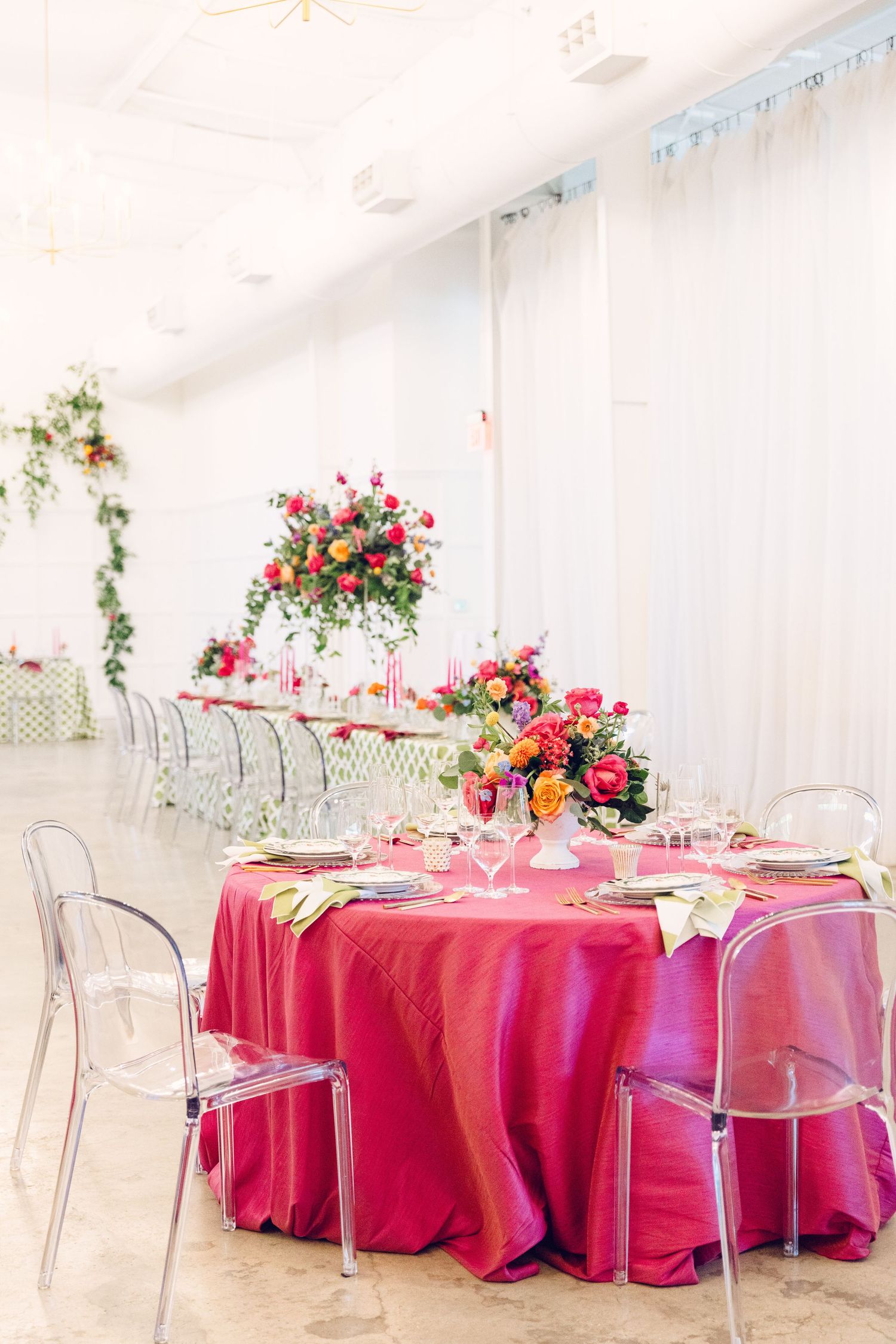 Elegant wedding reception table with hot pink linens, clear ghost chairs, and vibrant floral centerpieces in a white room.