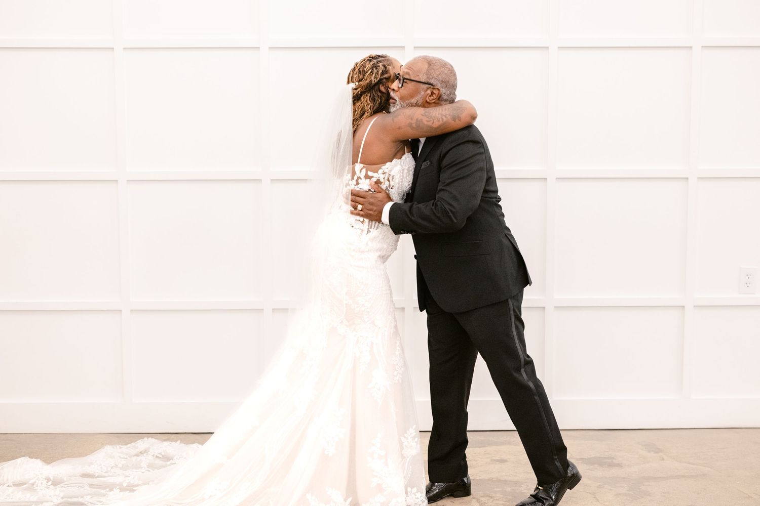 A bride and groom embrace in a romantic moment against a white wall on their wedding day.