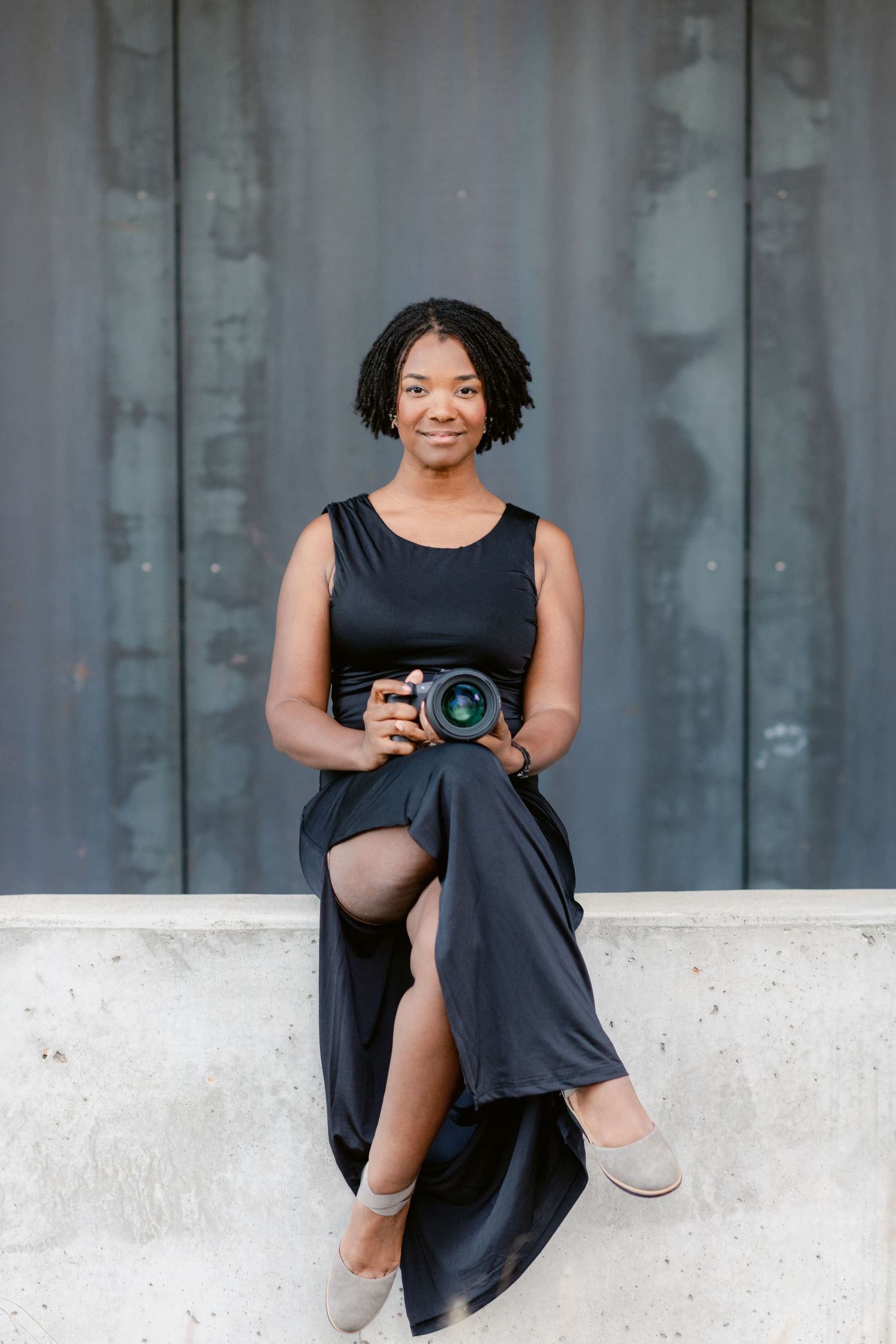 Photographer in black dress sits casually on white concrete ledge while holding professional camera.