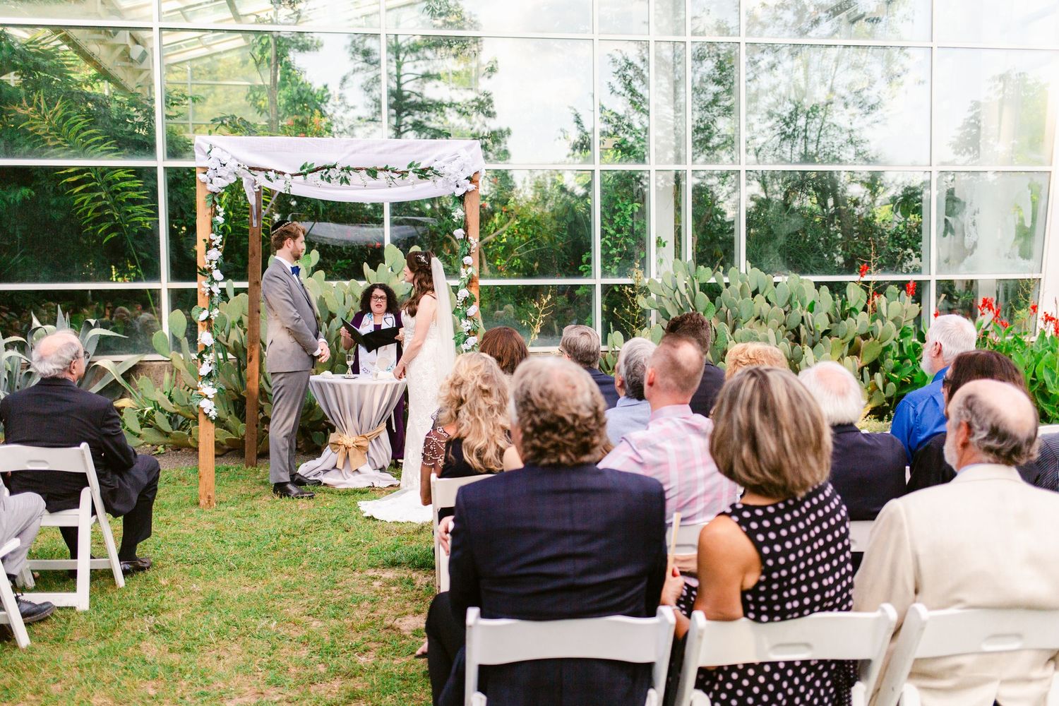 Outdoor wedding ceremony setup with white chairs and floral arrangements in garden setting.