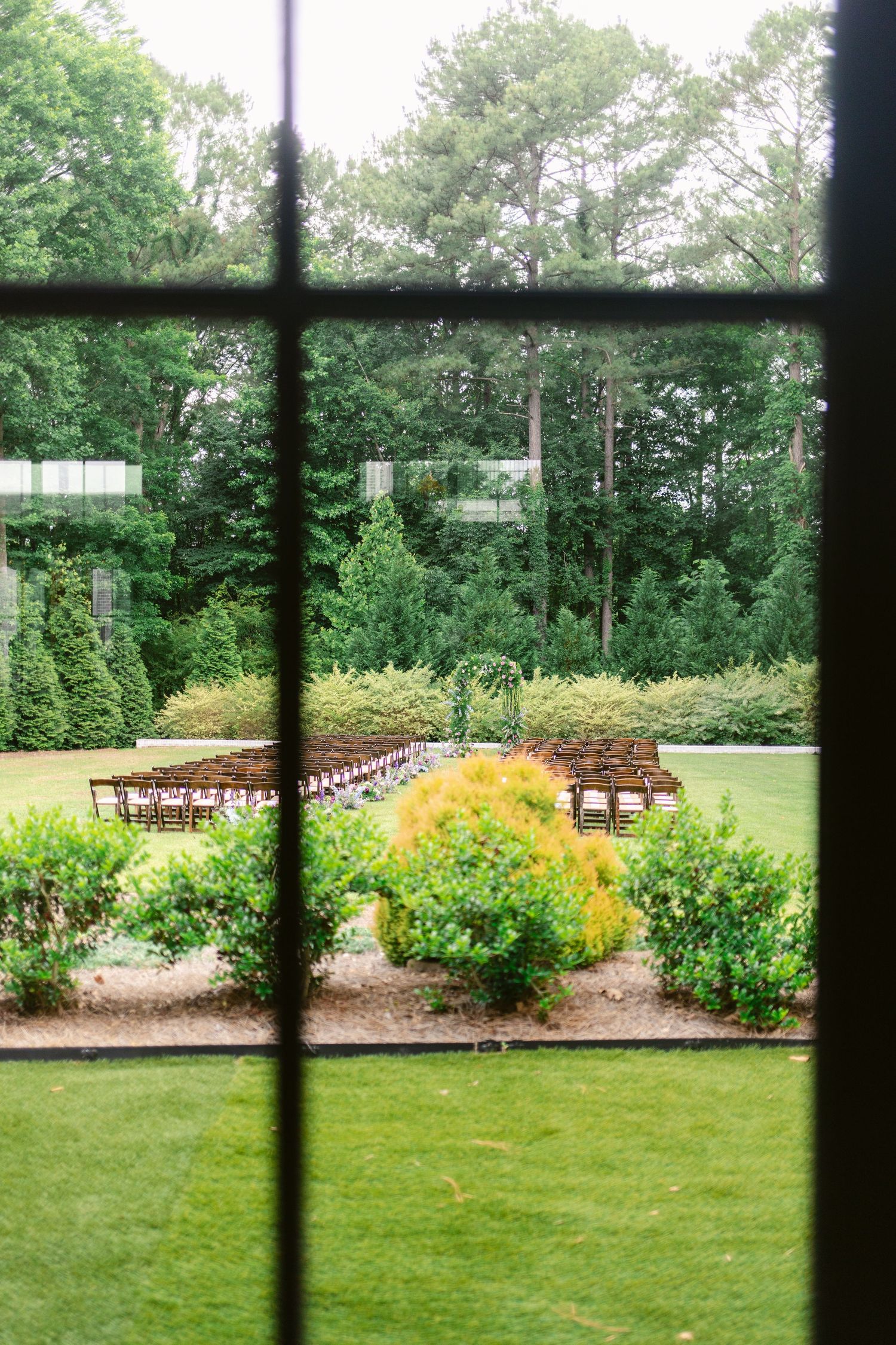 View through window panes of an outdoor garden wedding ceremony setup with wooden chairs and lush landscaping.