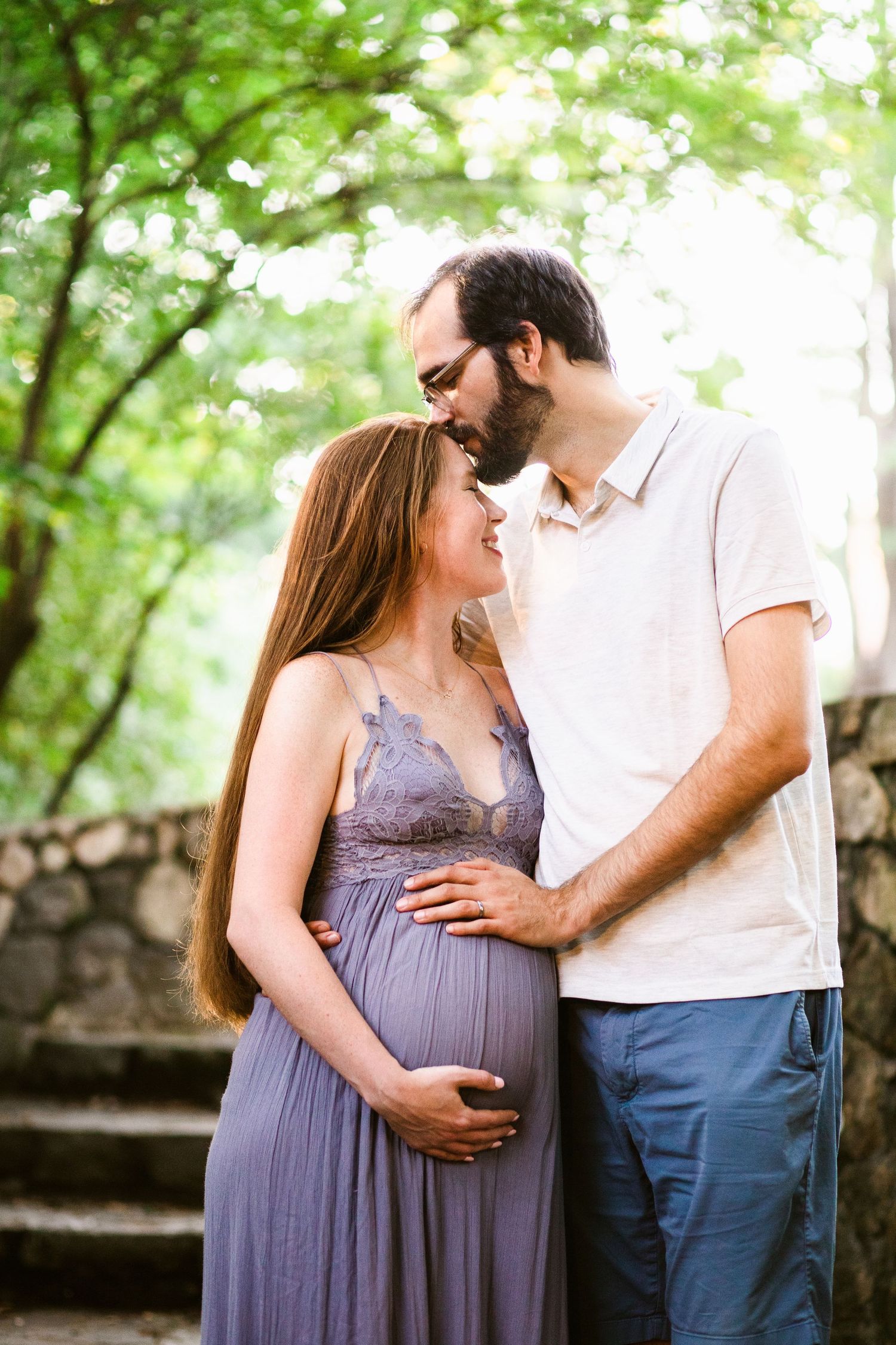 Maternity photo of couple in purple dress and white shirt among green trees.