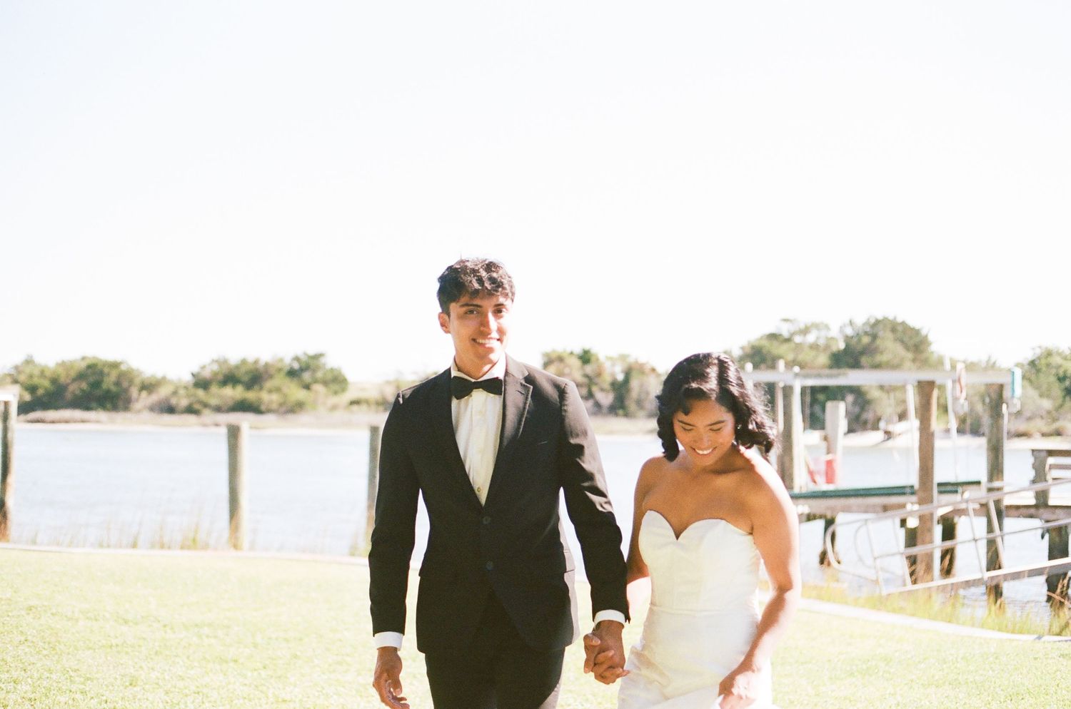 A happy couple walks hand in hand along a wooden pier by the water on their wedding day.