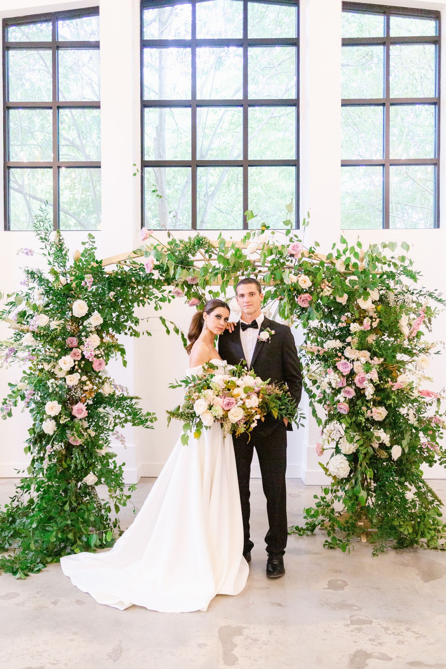 A couple stands under a lush floral arch with pink roses and greenery in an elegant indoor wedding setting.