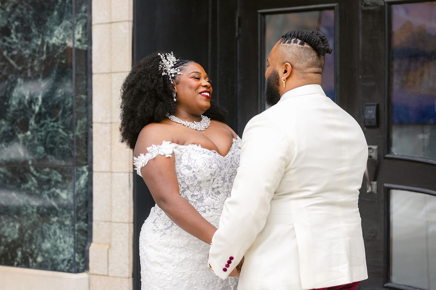 A plus-size bride in a lace off-shoulder gown holds hands with her groom in a white suit during their wedding day.