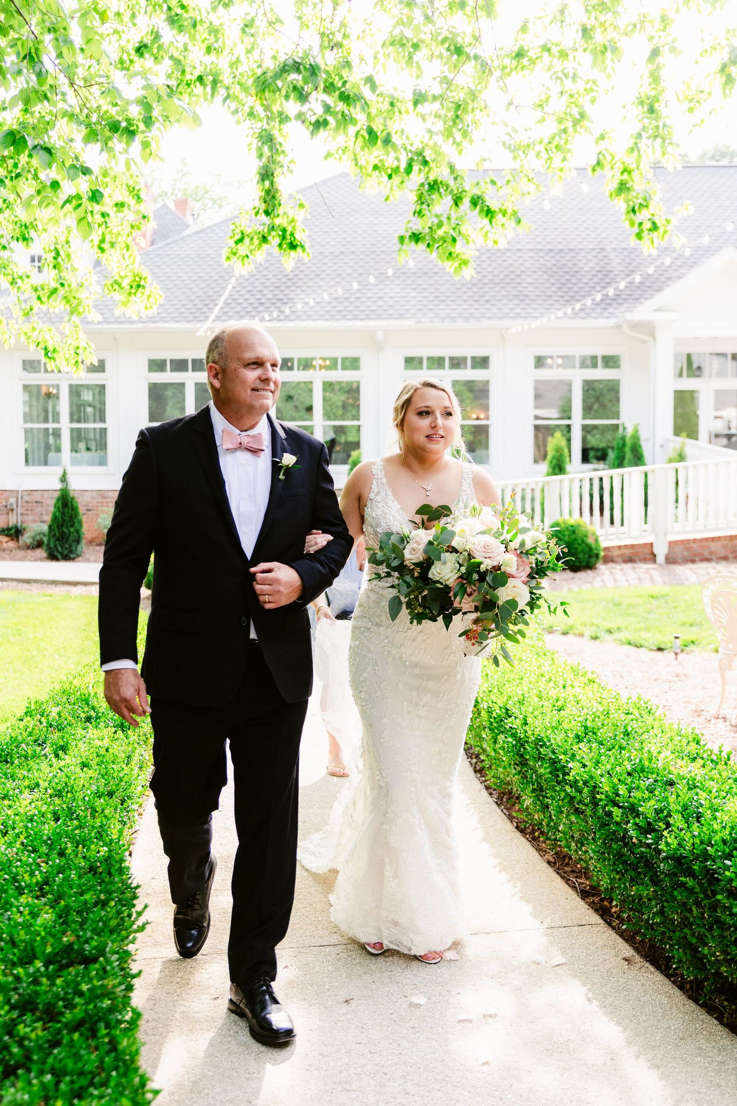 Wedding party walks down a covered outdoor walkway surrounded by lush greenery and white columns.