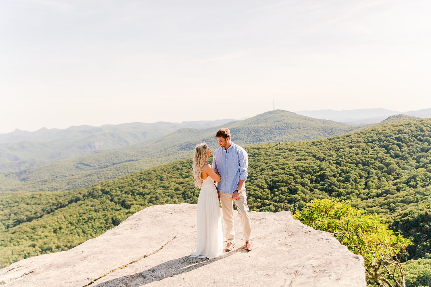 A couple shares a romantic moment on a mountain overlook surrounded by lush green forest views at sunset.