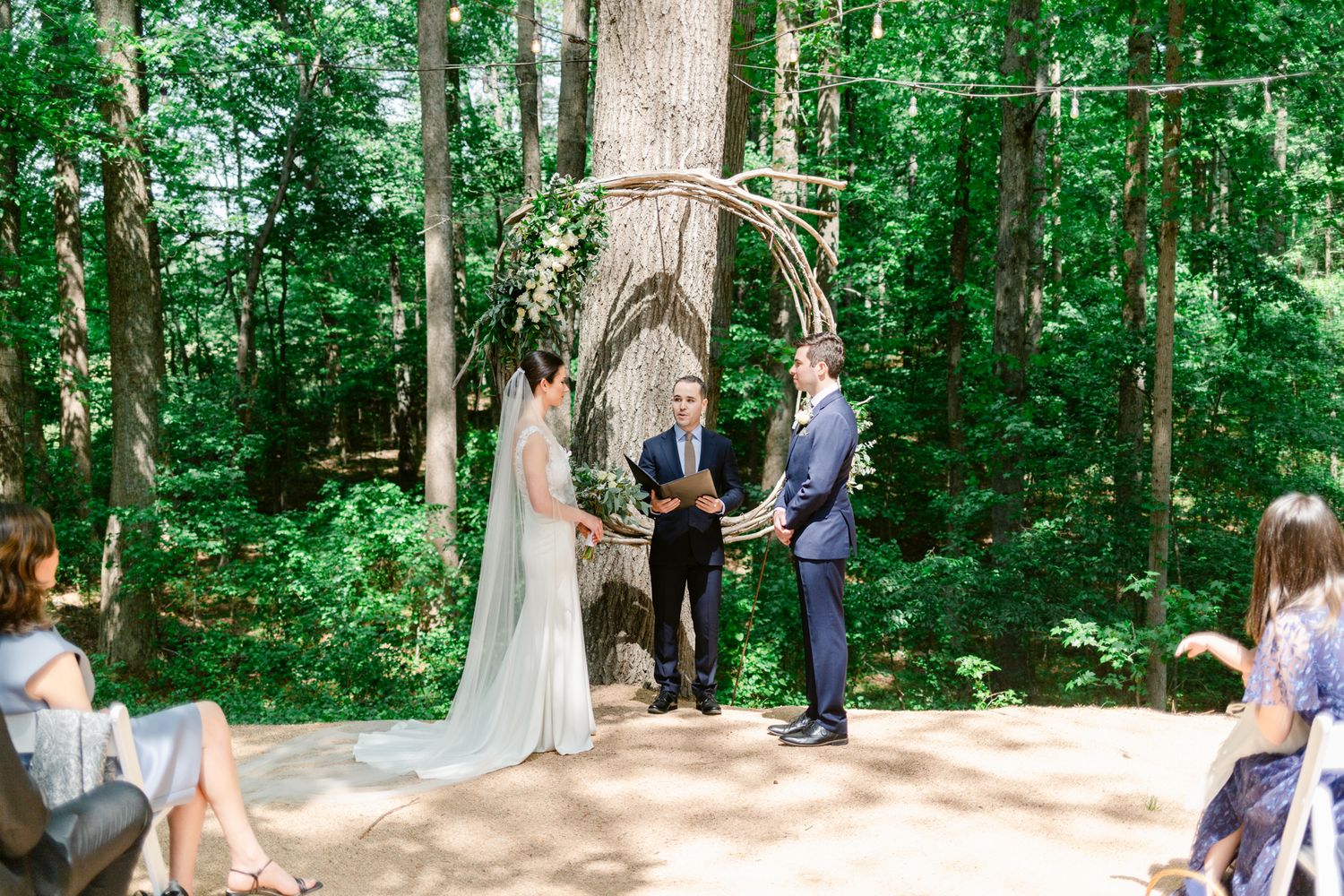 Wedding ceremony taking place in a sunlit forest clearing with guests gathered around white pillars.