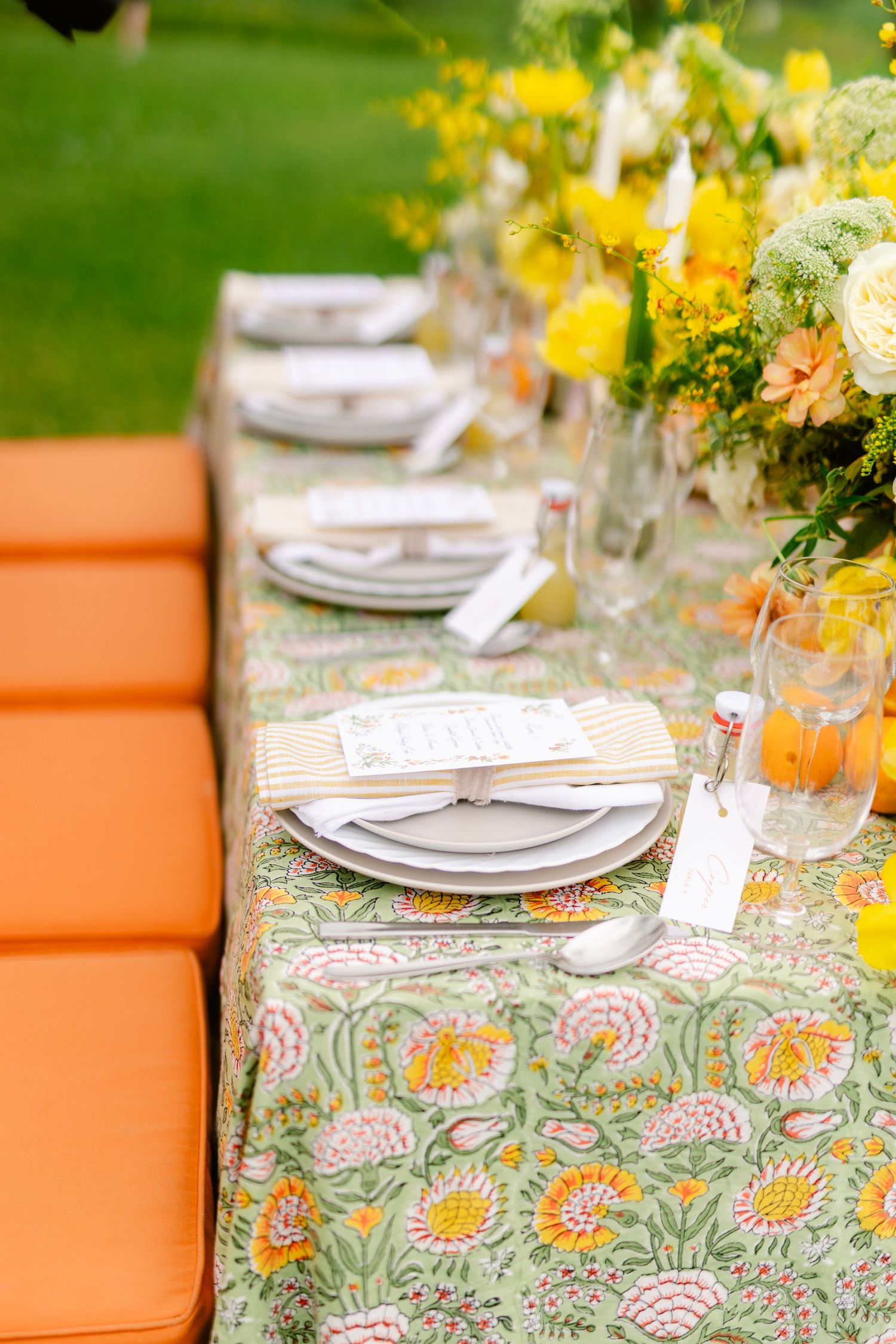 Elegant table setting with floral tablecloth, white plates, and orange seating against a green outdoor backdrop.