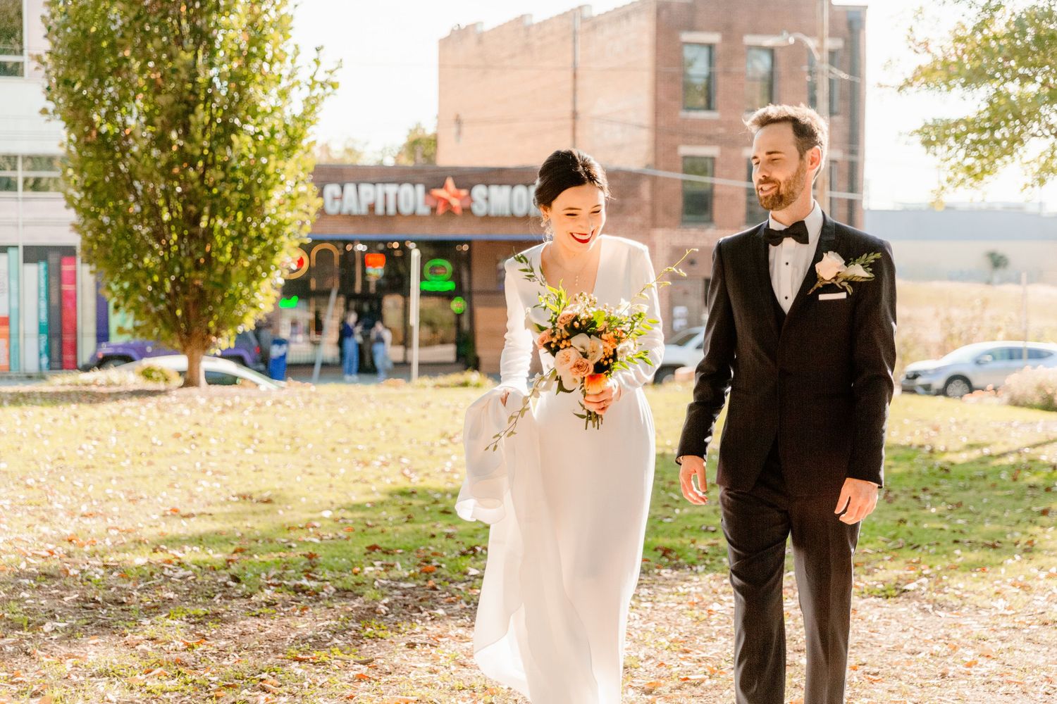 Bride and groom walk together through a park during autumn with fallen leaves scattered on the ground.