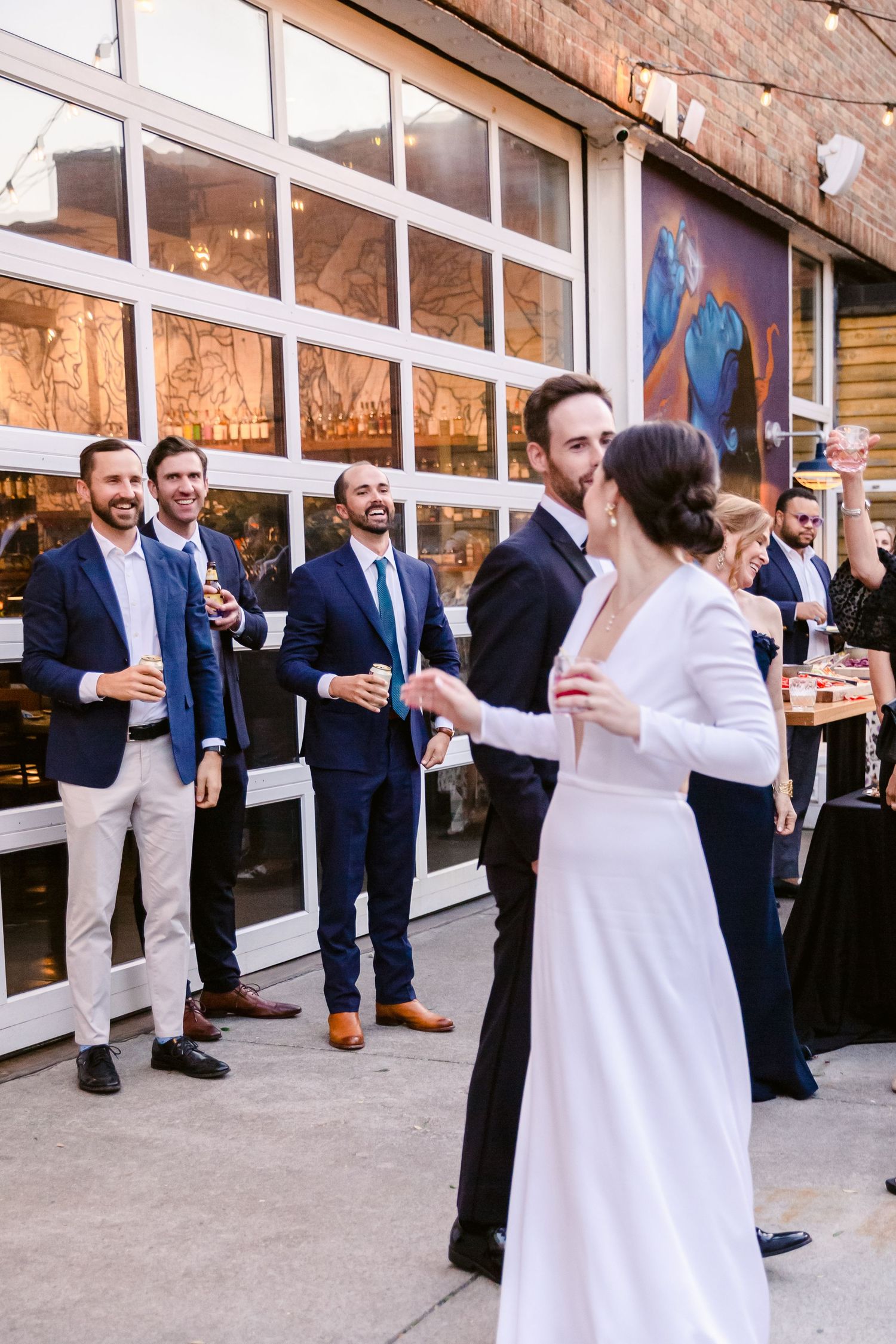 Wedding guests in navy blazers gather outside a venue with large windows during a celebration.