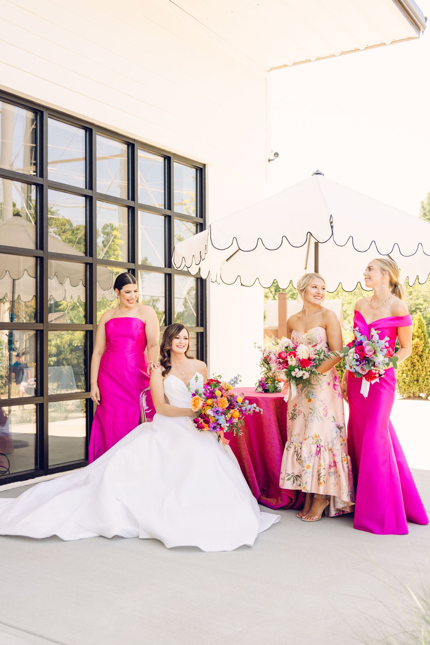Wedding party in bright pink dresses and white gown posed in front of modern windows in sunlit venue.