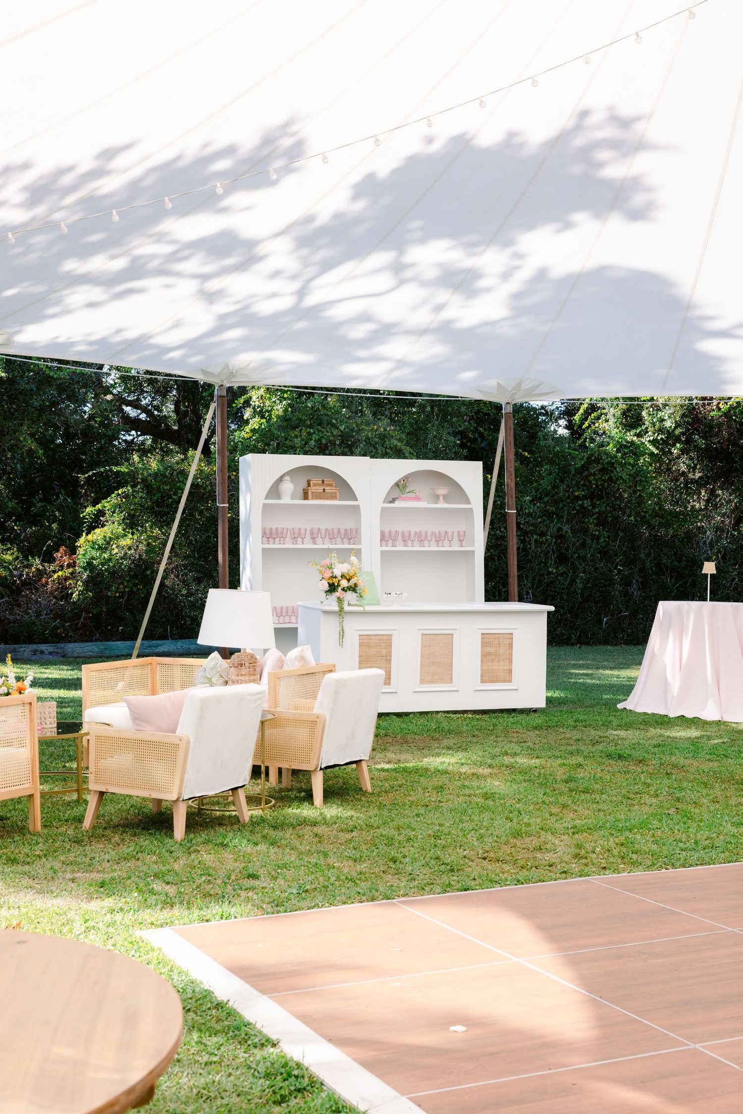 Elegant outdoor wedding setup with white hutch display, wicker chairs and dance floor under a white tent canopy.