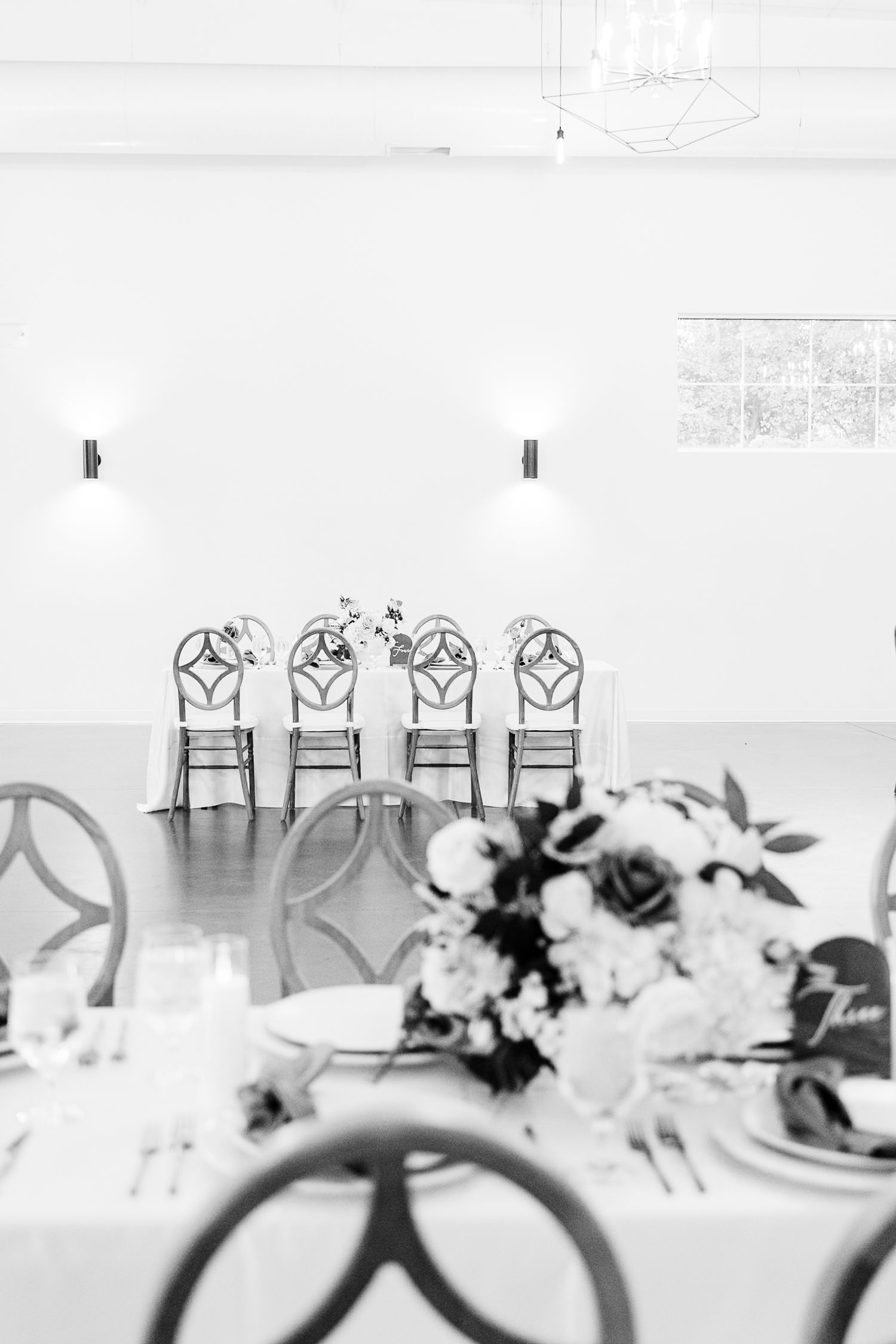 Wedding reception tables decorated with elegant white linens and floral centerpieces feature ornate high-back chairs.
