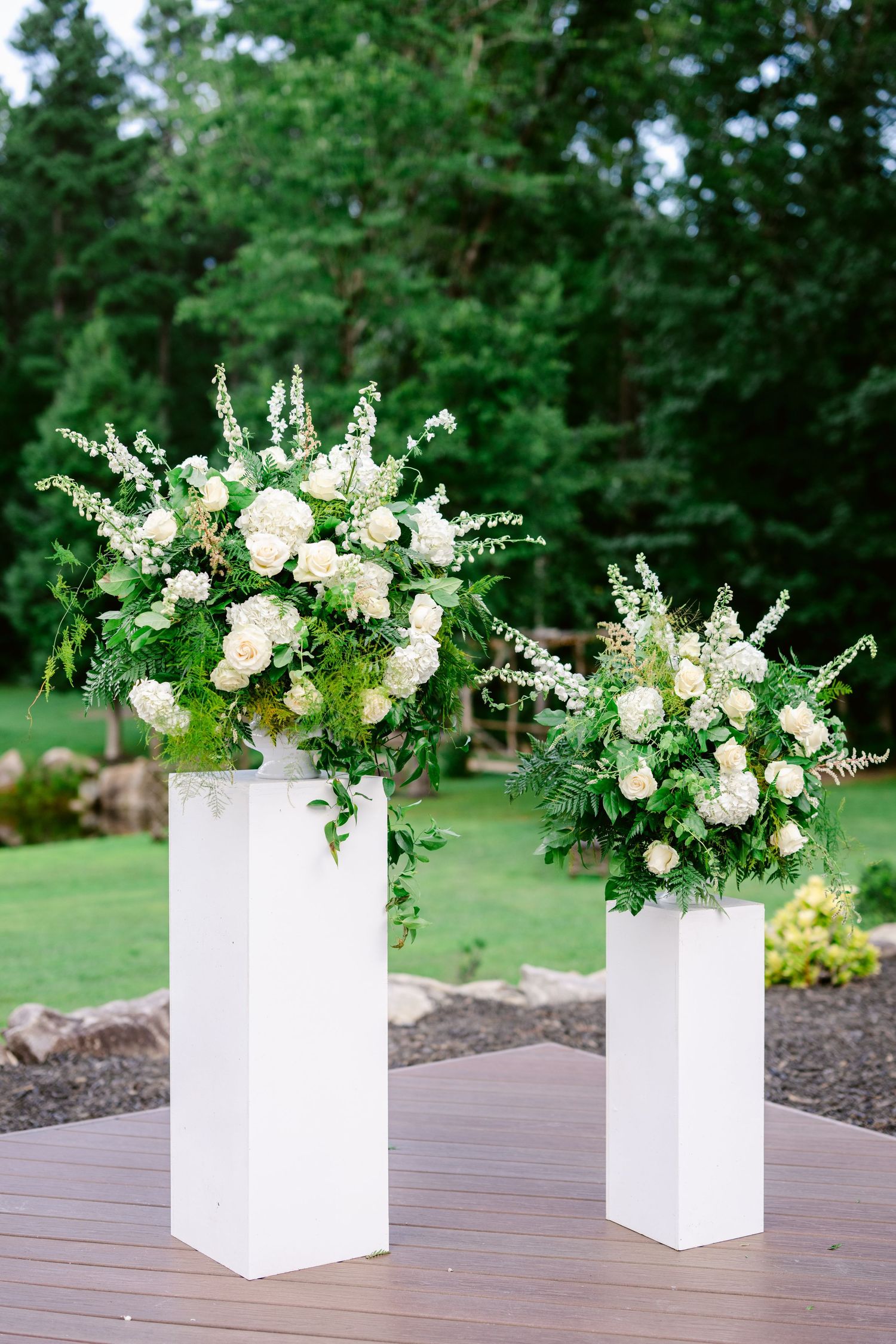 Four elegant white floral arrangements in matching white rectangular vases displayed on a wooden deck with green lawn background.