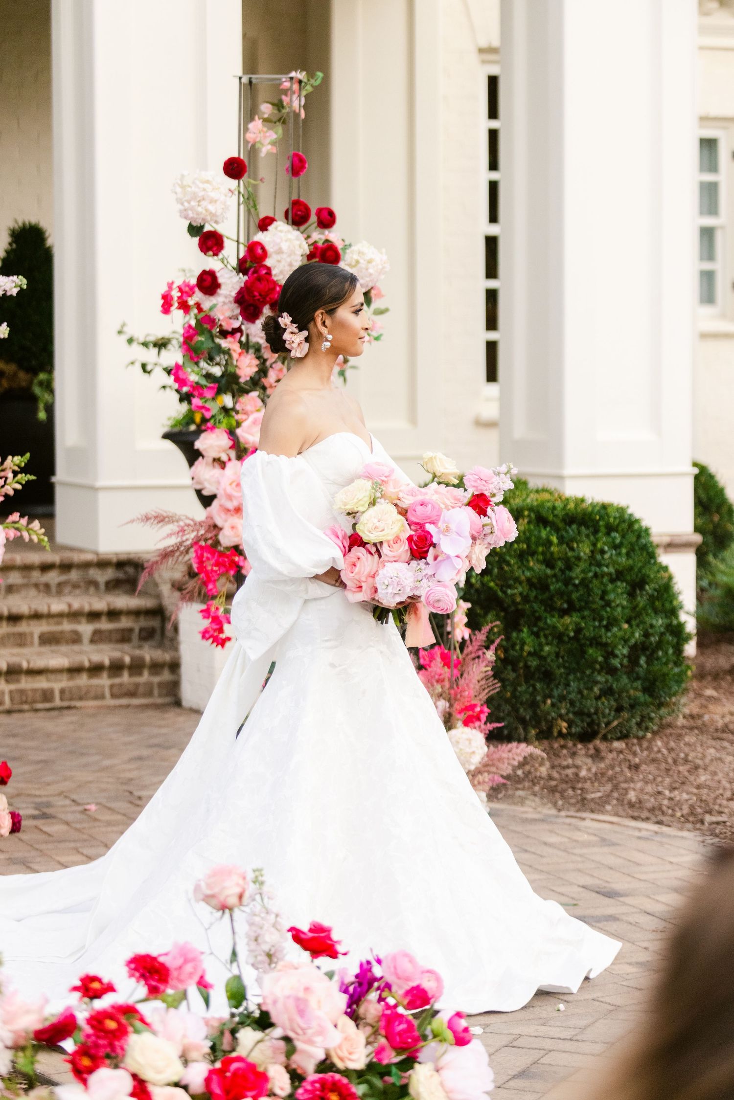 Bride in flowing white gown photographed with pink floral arrangements at elegant venue.