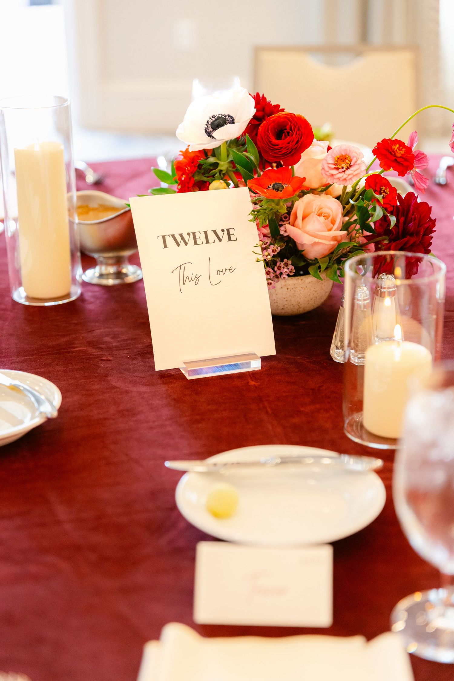 Table setting with red and pink floral centerpiece, place cards, and candles on burgundy tablecloth.