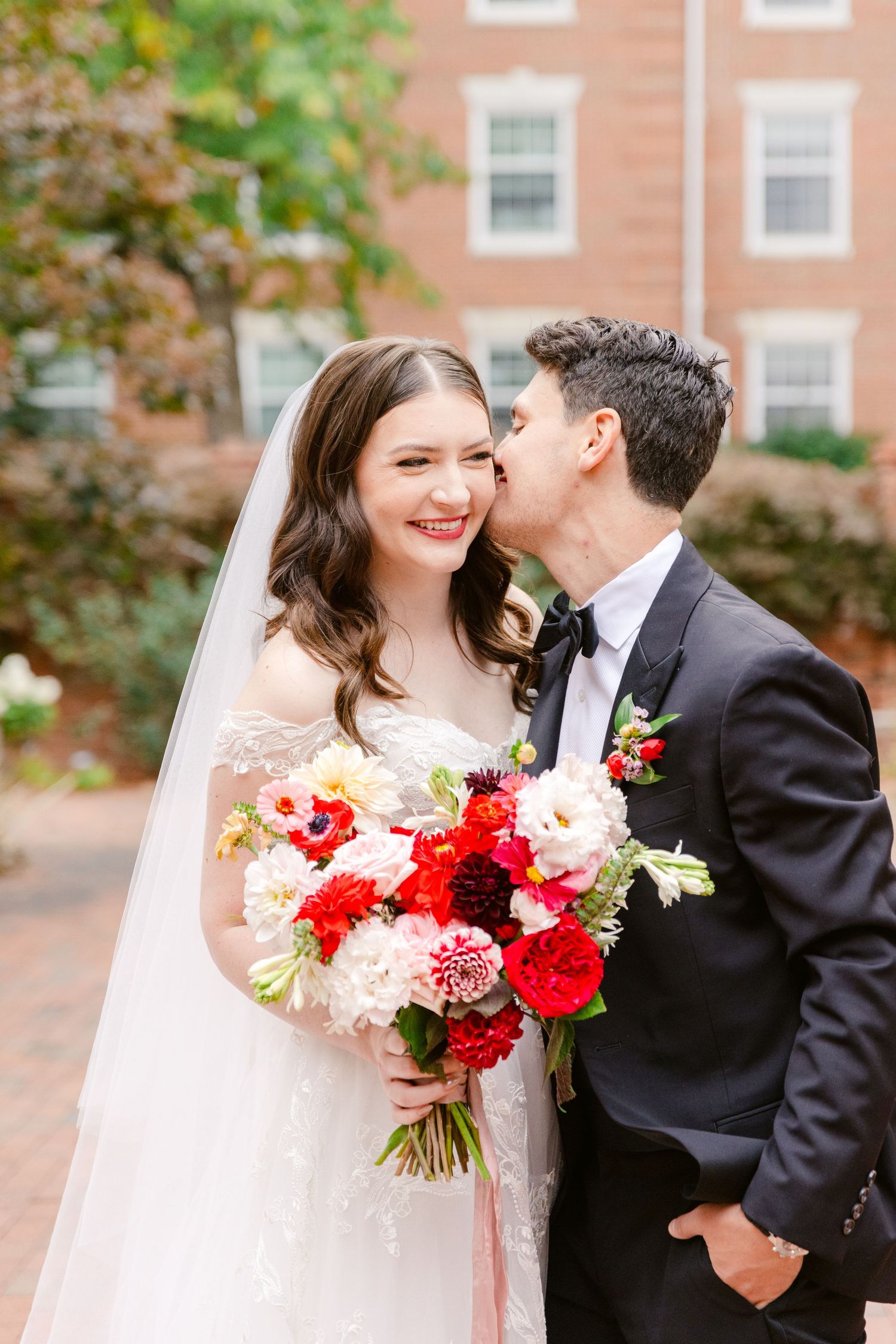 Bride smiling while groom kisses her cheek, holding vibrant red and pink floral bouquet outdoors near brick building.